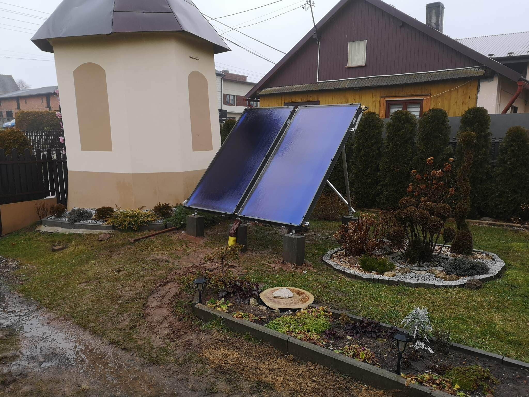 Dwa panele słoneczne zamontowane na betonowych podstawach w przydomowym ogrodzie, obok kapliczki i domu z drewnianą elewacją. Na panelach widoczne krople wody, a ziemia jest błotnista.