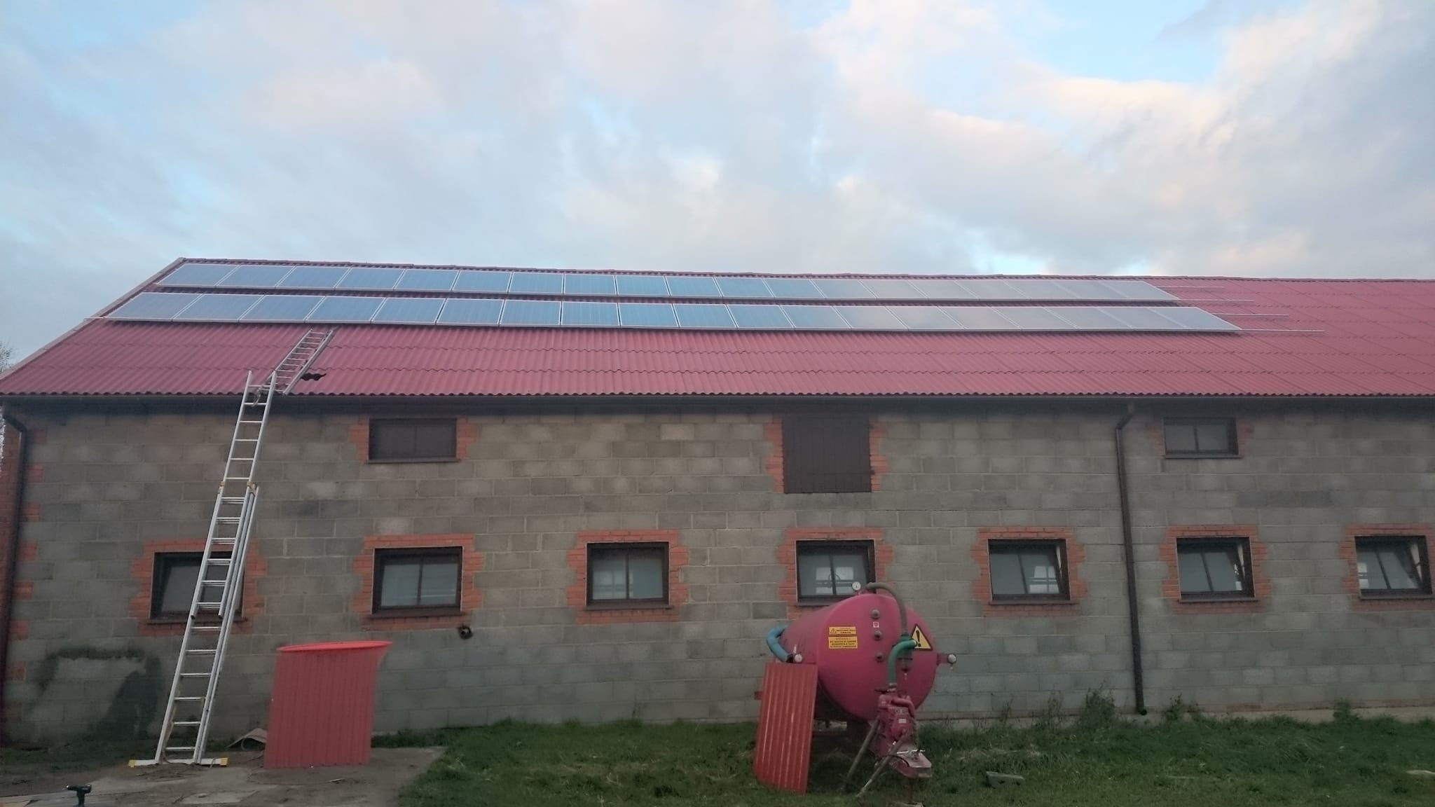 Panele fotowoltaiczne na czerwonym dachu budynku gospodarczego z szarego betonu, drabina oparta o dach, czerwony zbiornik na podwórzu, pochmurne niebo.