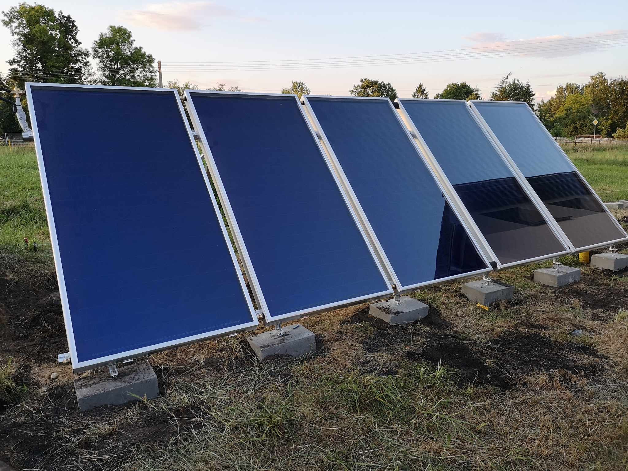 Rząd pięciu paneli słonecznych ustawionych na betonowych blokach na trawiastym terenie, widoczne odbicia nieba na powierzchni paneli.