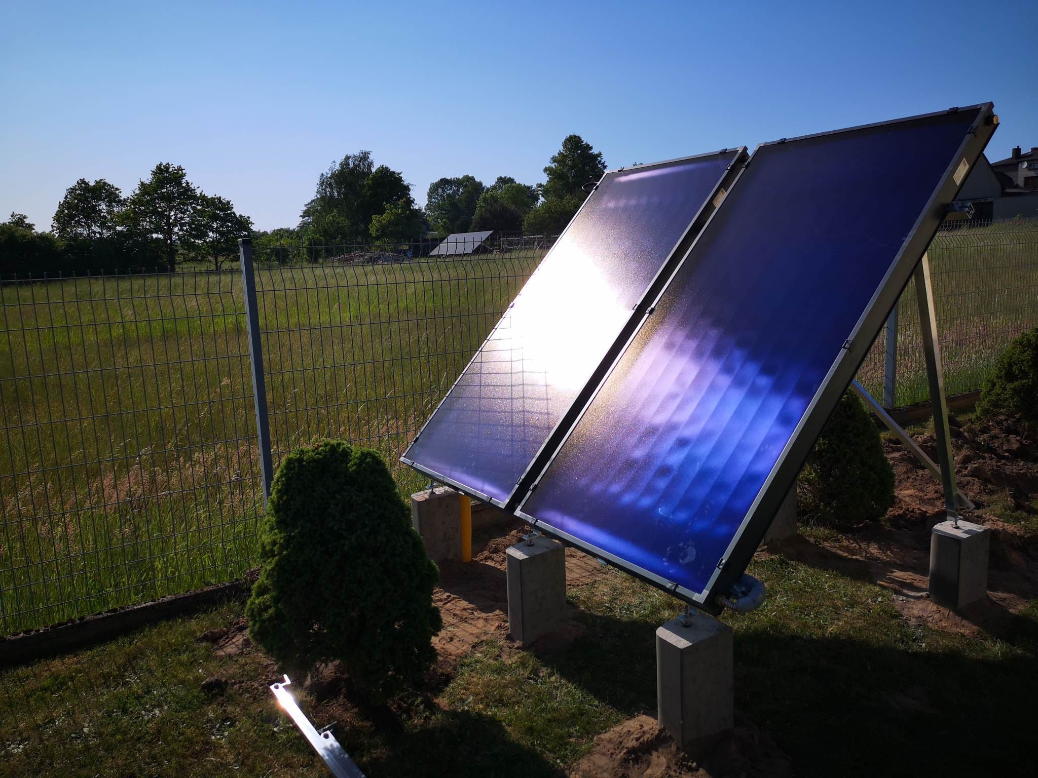 Dwa kolektory słoneczne zamontowane na betonowych podstawach w ogrodzie, z widocznym odbiciem słońca na powierzchni paneli i ogrodzeniem w tle.