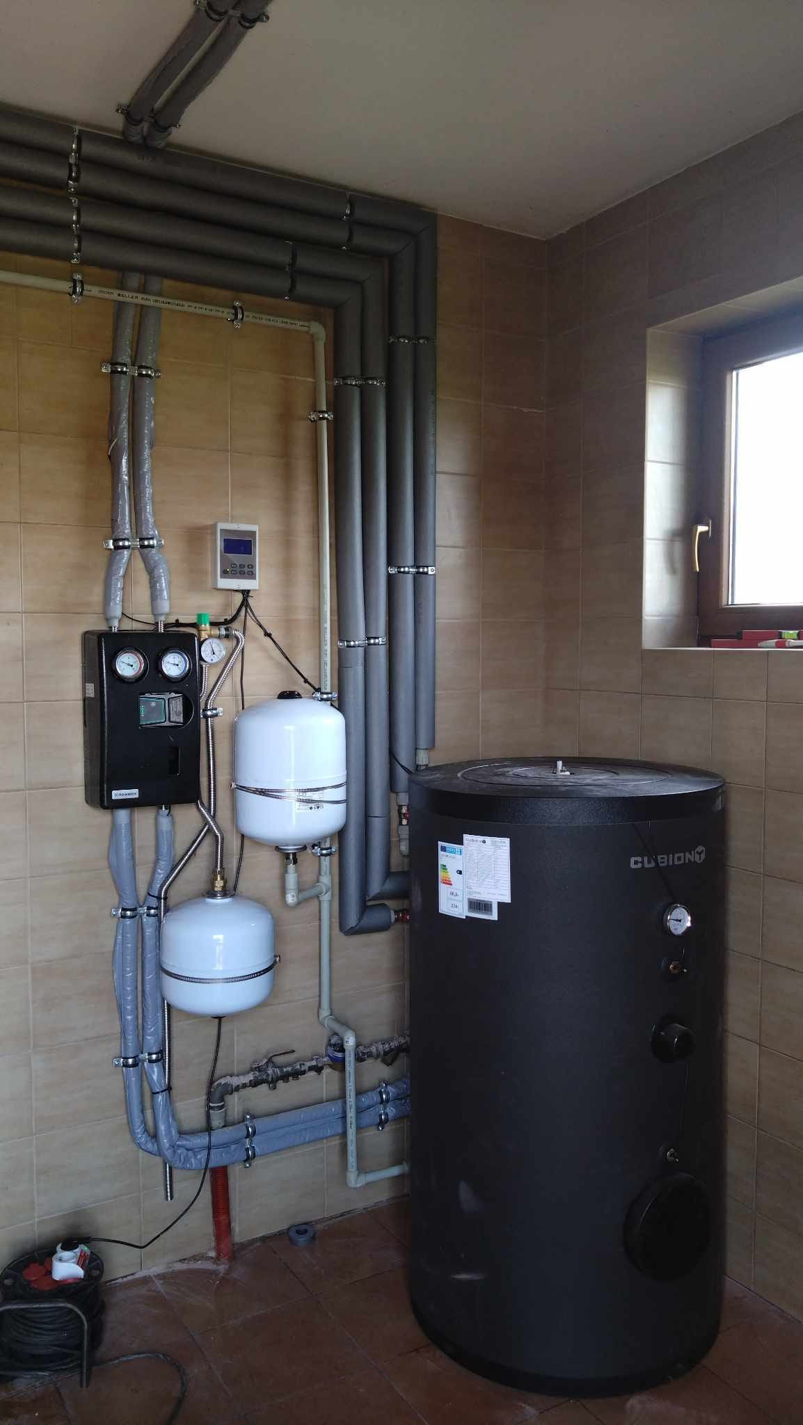 Instalacja kotłowni z czarnym zbiornikiem akumulacyjnym, dwoma białymi zbiornikami wyrównawczymi, sterownikiem i rurami w izolacji, widok z bliska na brązowe płytki podłogowe i ścienne.