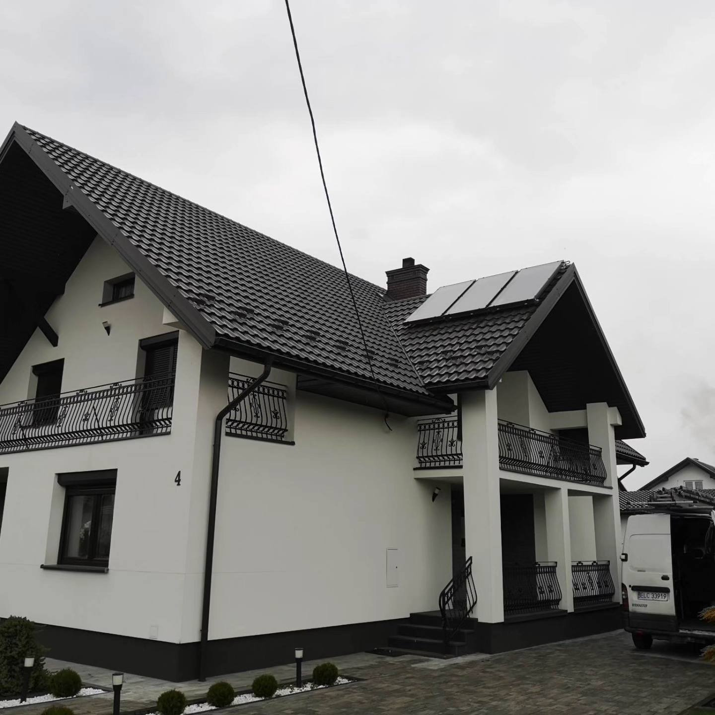 Nowoczesny dom jednorodzinny z białymi ścianami, czarnym dachem z dachówki i trzema panelami na dachu, widok z boku, z zaparkowanym białym vanem z otwartymi drzwiami w tle.