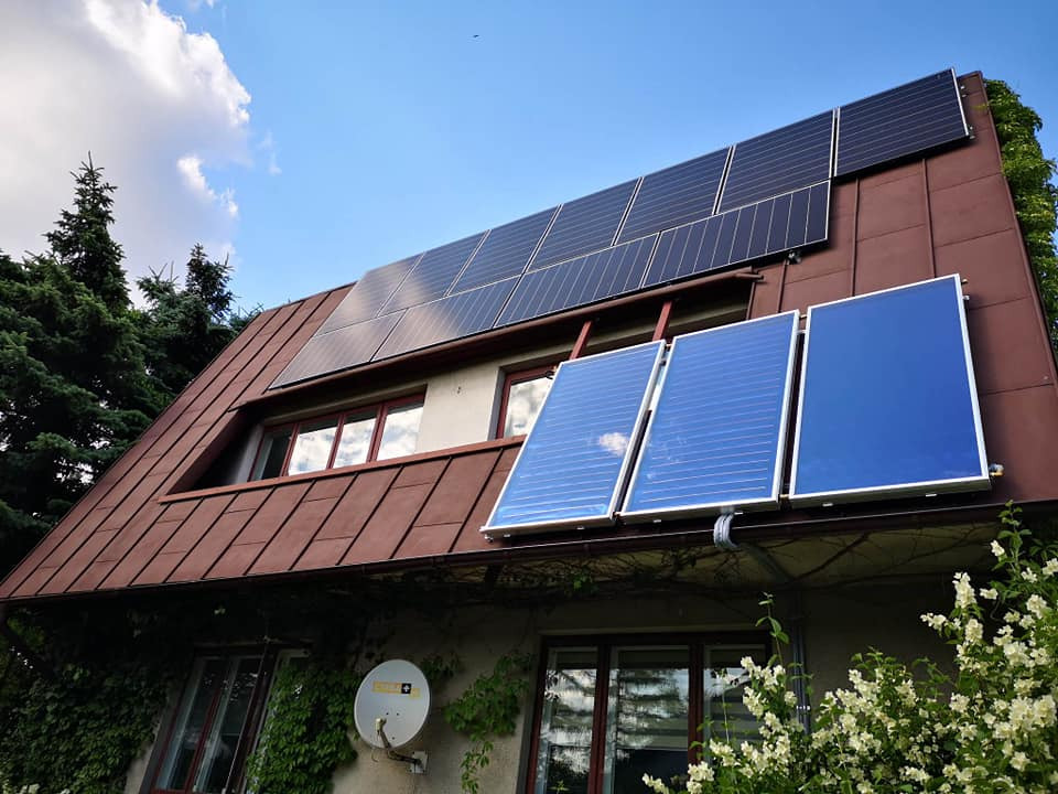 Dom z brązowym dachem, na którym zamontowano panele fotowoltaiczne i termiczne, widoczne z dołu, z krzewami kwitnącymi na pierwszym planie.