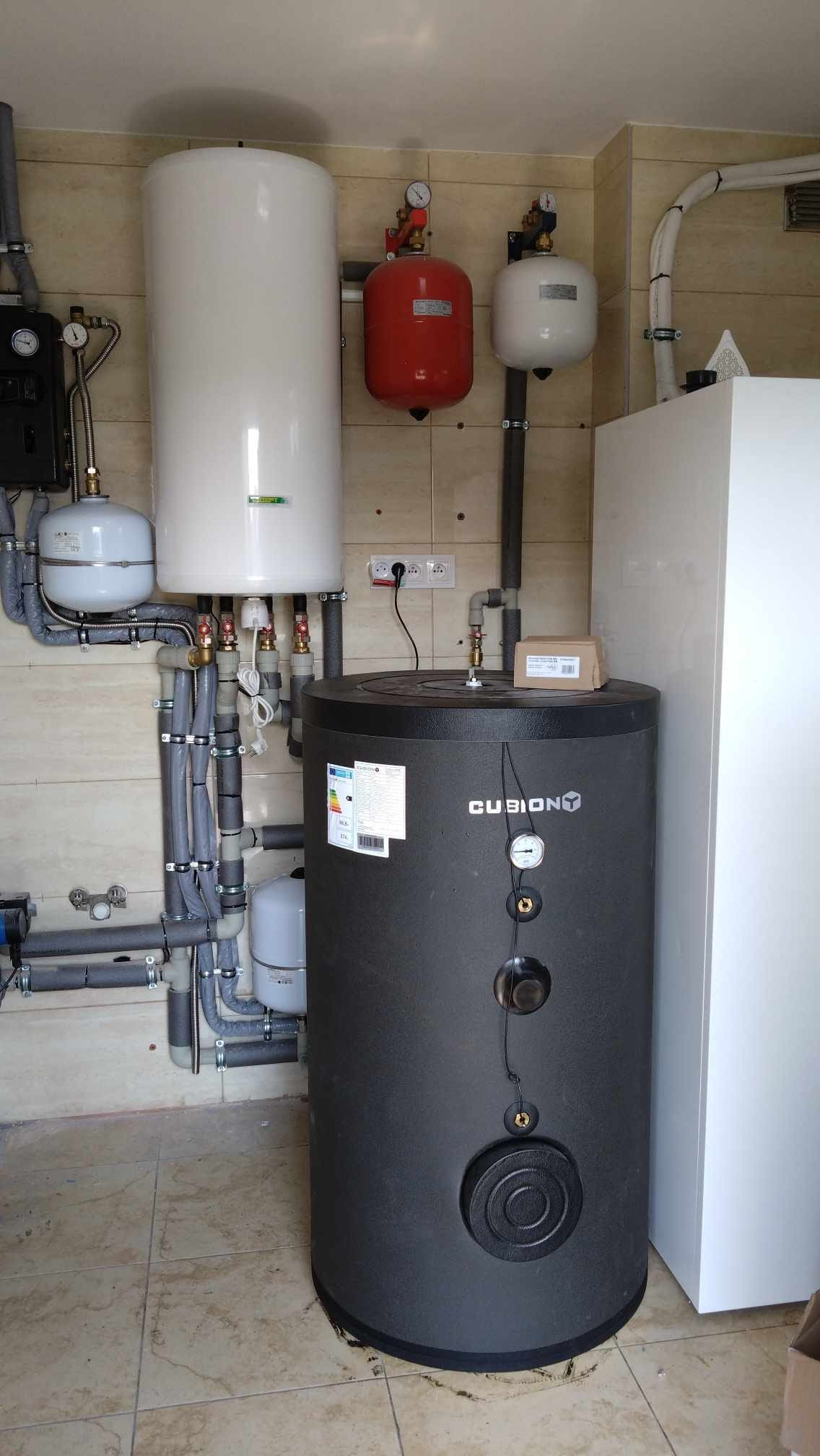 Instalacja z czarnym zbiornikiem buforowym Cubiony, białym bojlerem, czerwonym naczyniem wzbiorczym i szarą izolacją rur na beżowej ścianie z płytek.
