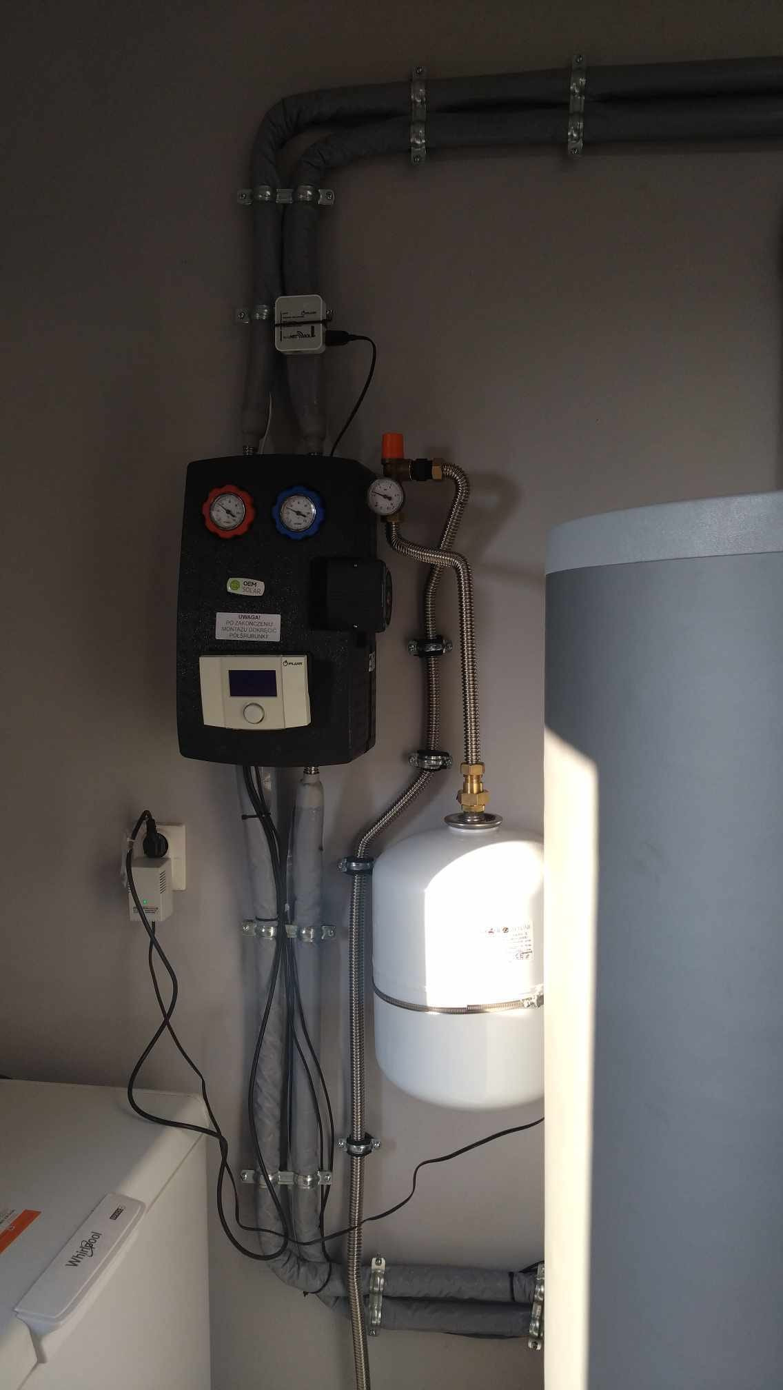 Zamontowany system kolektorów słonecznych z czarnym panelem sterowania, białym zbiornikiem wyrównawczym i szarym zasobnikiem wody, rury izolowane szarą otuliną.