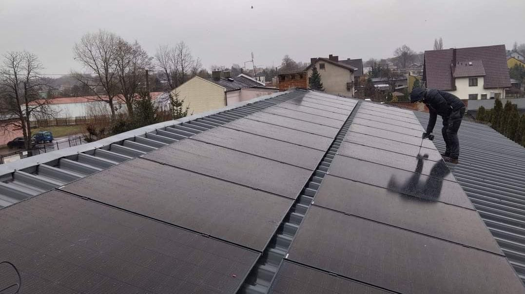 Montaż paneli słonecznych na metalowym dachu, widoczne krople deszczu i sylwetka instalatora w ciemnej odzieży.