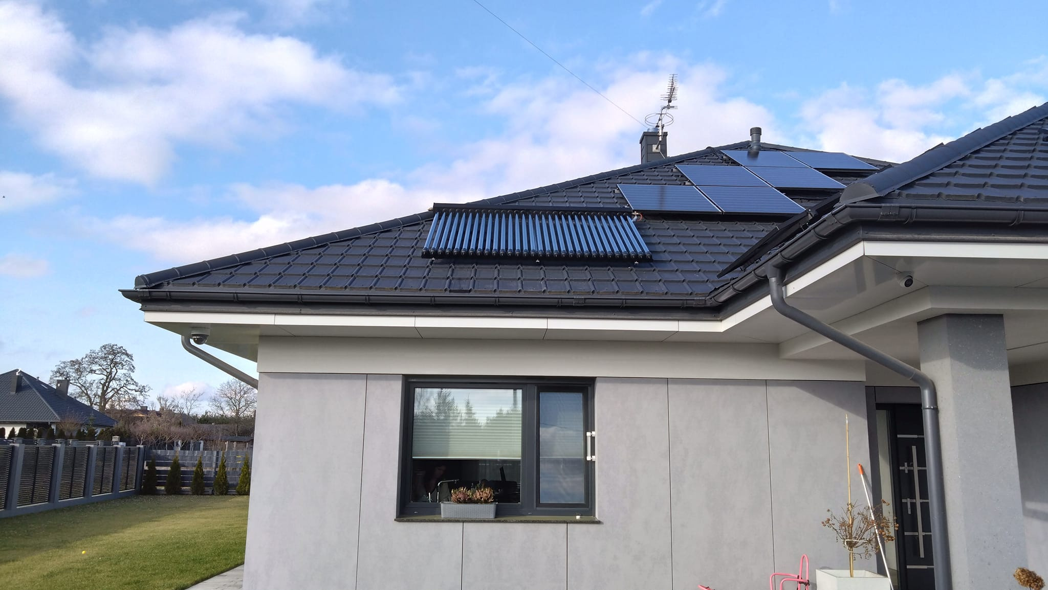 Rząd rur próżniowych i panele fotowoltaiczne na ciemnym, skośnym dachu nowoczesnego domu z szarej elewacji w słoneczny dzień.