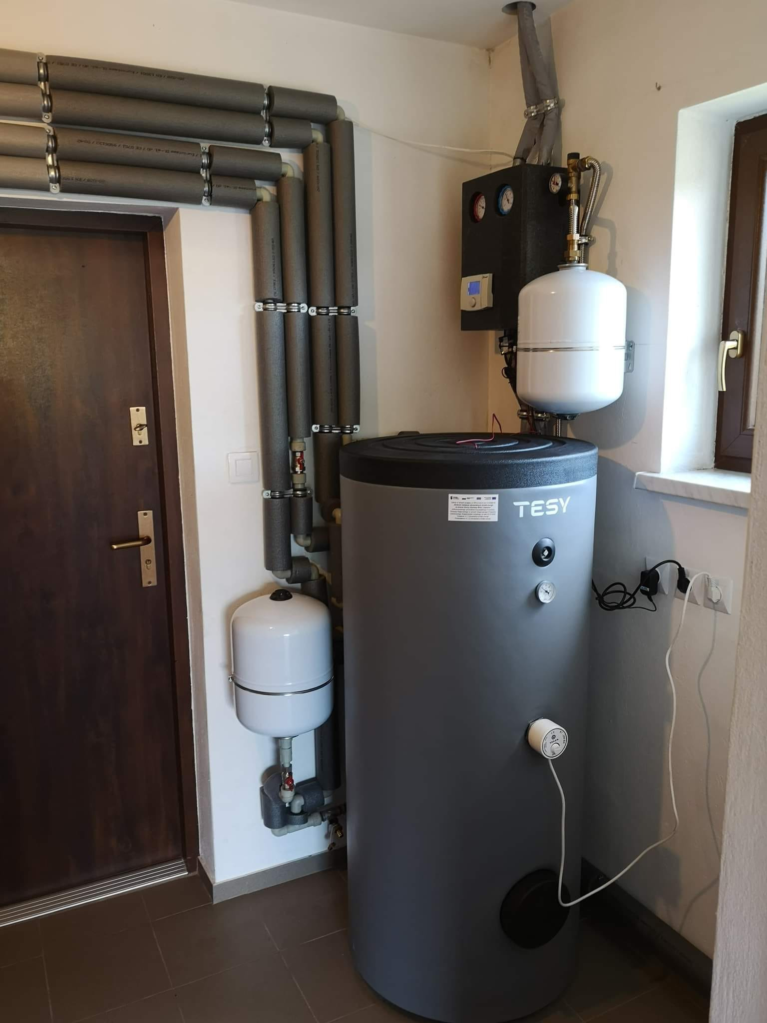Szary zasobnik ciepłej wody użytkowej marki TESY z podłączonymi rurami w izolacji termicznej, zamontowany w rogu pomieszczenia obok drewnianych drzwi i okna.