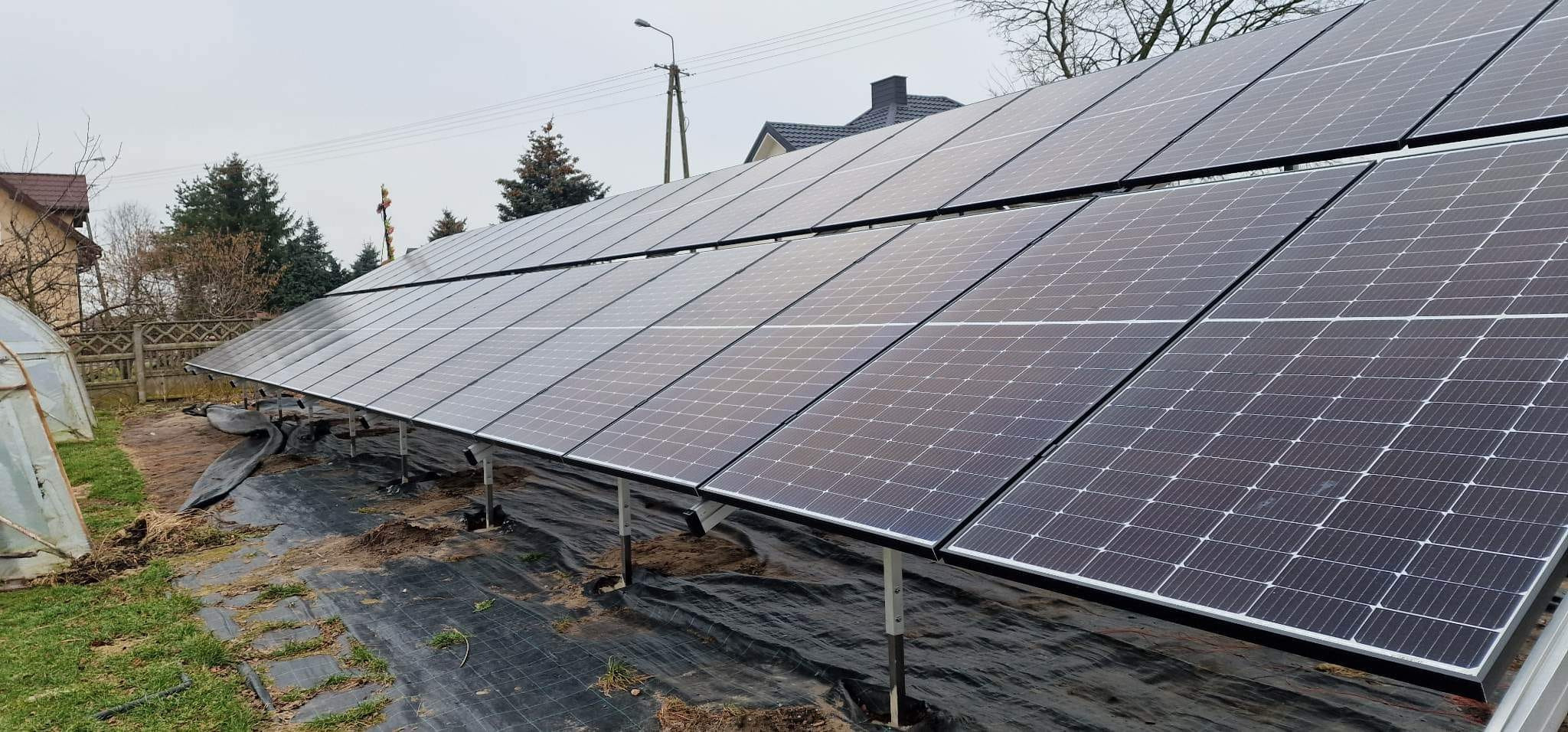 Rząd paneli fotowoltaicznych na gruncie, zamontowanych na metalowych wspornikach, z widoczną czarną agrowłókniną pod spodem i fragmentem szklarni w tle.