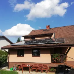 Dom jednorodzinny z brązowym dachem i panelami słonecznymi na dachu, widok z ogrodu z meblami ogrodowymi i kwiatami w skrzynkach okiennych, błękitne niebo z białymi chmurami.