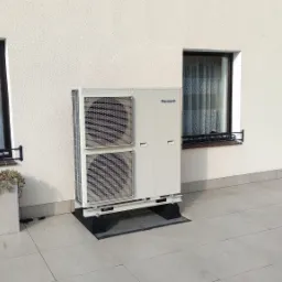 Biała pompa ciepła marki Panasonic zamontowana na zewnątrz budynku, widoczna na tle białej ściany z oknami i balkonem z ozdobną balustradą.