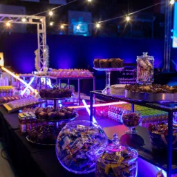 Stół z przekąskami na imprezie firmowej, podświetlony neonami i girlandami żarówek, z muffinami, słodyczami w słoikach i napojami w tle niebieski ekran z napisem 'Witaj!'