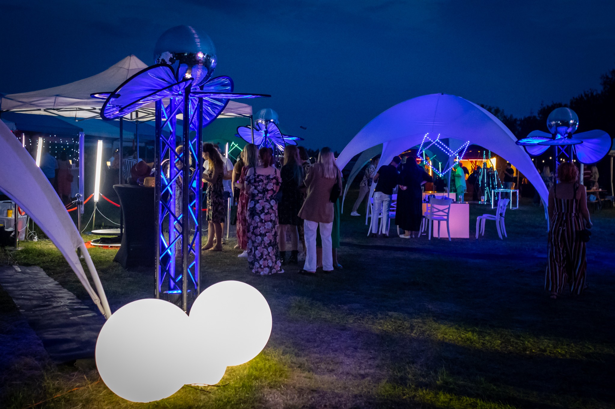 Wieczorny plener z oświetlonymi namiotami i dekoracjami, grupa osób w tle, oświetlenie ledowe w kolorze niebieskim i białym.