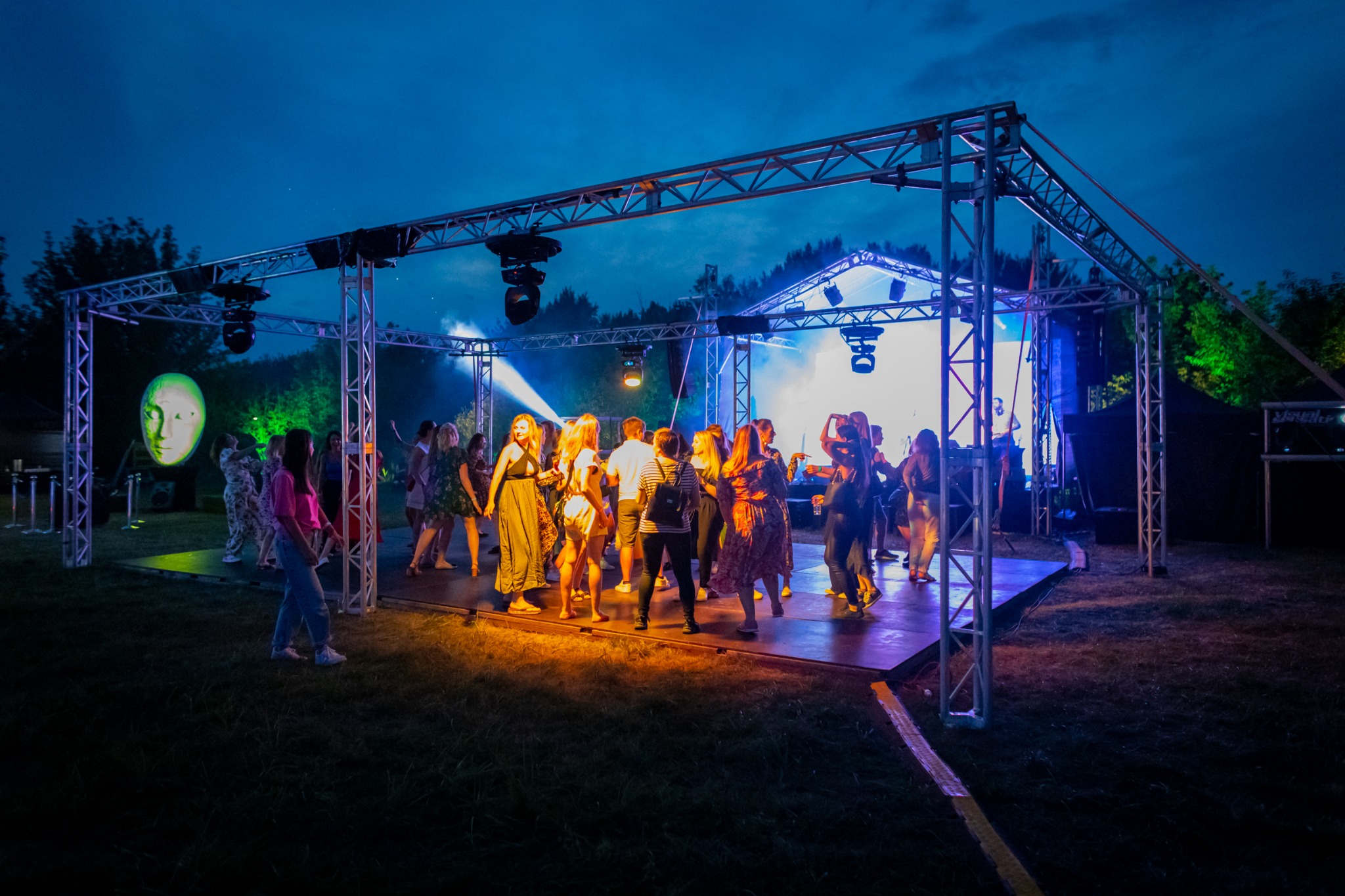 Grupa osób tańczących na podwyższeniu pod metalową konstrukcją oświetleniową podczas imprezy plenerowej o zmierzchu, z projekcją twarzy na ekranie w tle.