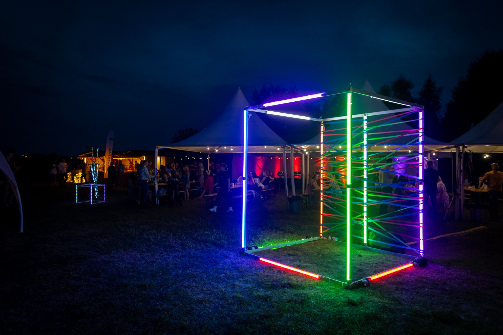 Oświetlona, kolorowa instalacja w kształcie sześcianu na trawiastym terenie podczas imprezy plenerowej, w tle widoczne namioty i bawiący się ludzie.