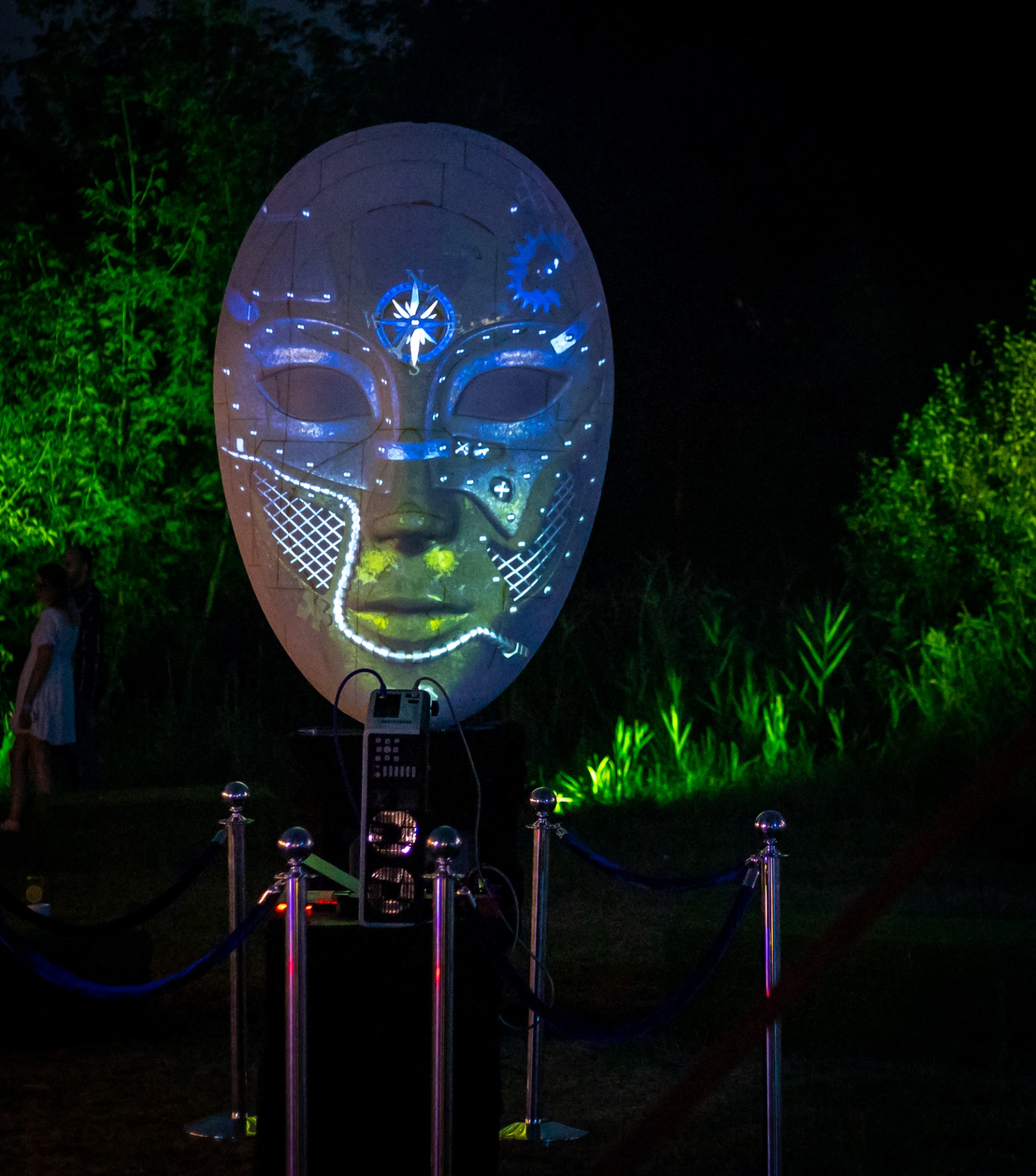 Projekcja świetlna na dużej masce w plenerze, oświetlenie dekoracyjne, barierki z aksamitną liną wyznaczają przestrzeń, w tle zieleń.