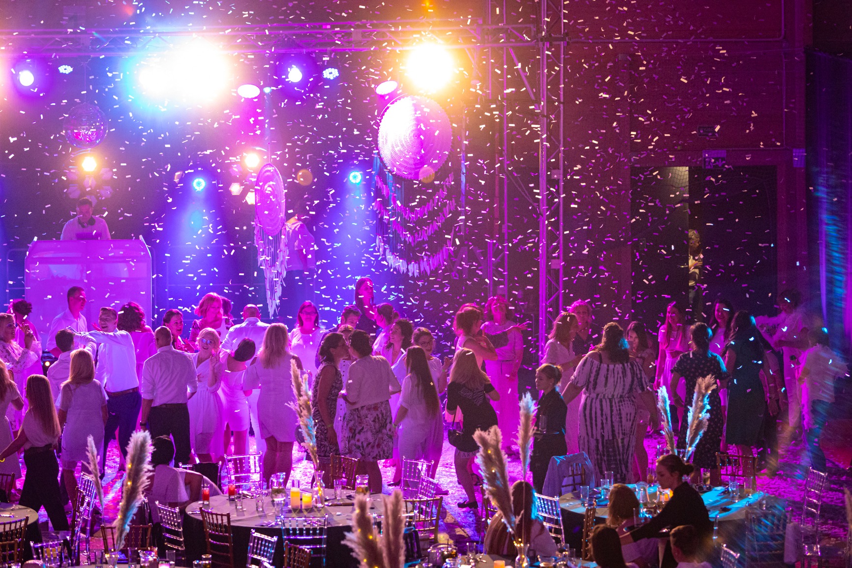Tłum ludzi tańczących pod światłami dyskotekowymi i konfetti, DJ na podwyższeniu w tle, dekoracje stołów w pierwszym planie.