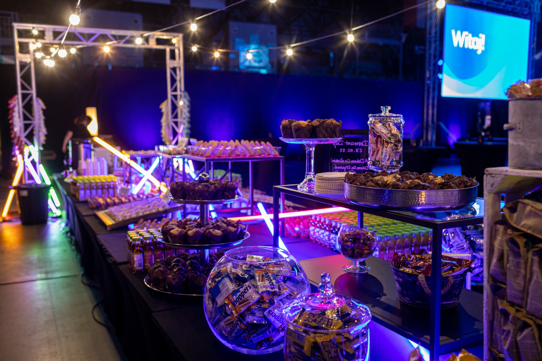 Stół z przekąskami na imprezie firmowej, podświetlony neonami i girlandami żarówek, z muffinami, słodyczami w słoikach i napojami w tle niebieski ekran z napisem 'Witaj!'