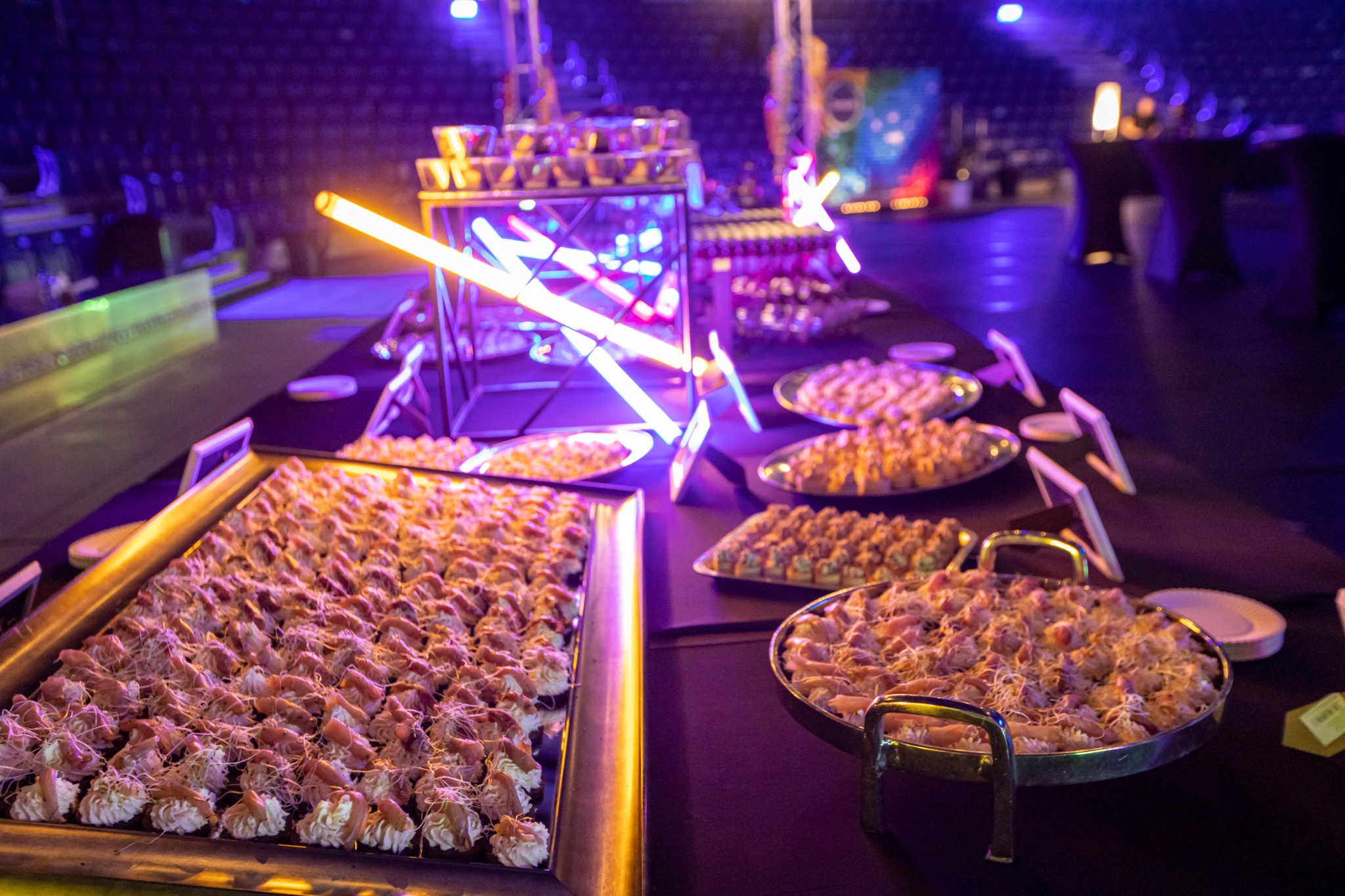 Stół cateringowy z przekąskami podświetlony neonowym światłem na evencie, w tle widoczne miejsca siedzące dla publiczności.