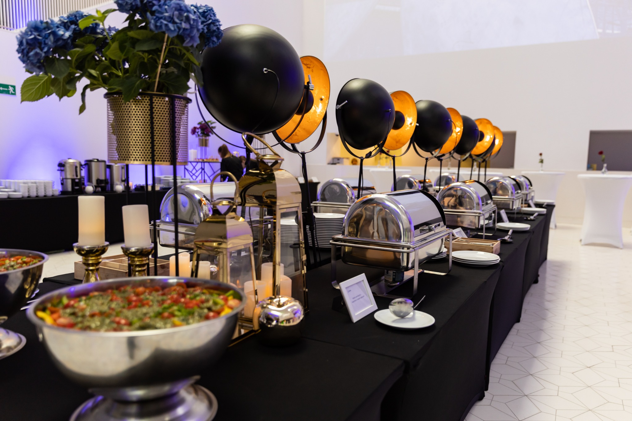 Szczeciński stół bufetowy na imprezie, z czarnym obrusem, metalowymi naczyniami do podgrzewania potraw, dekoracjami kwiatowymi i lampami w stylu retro.