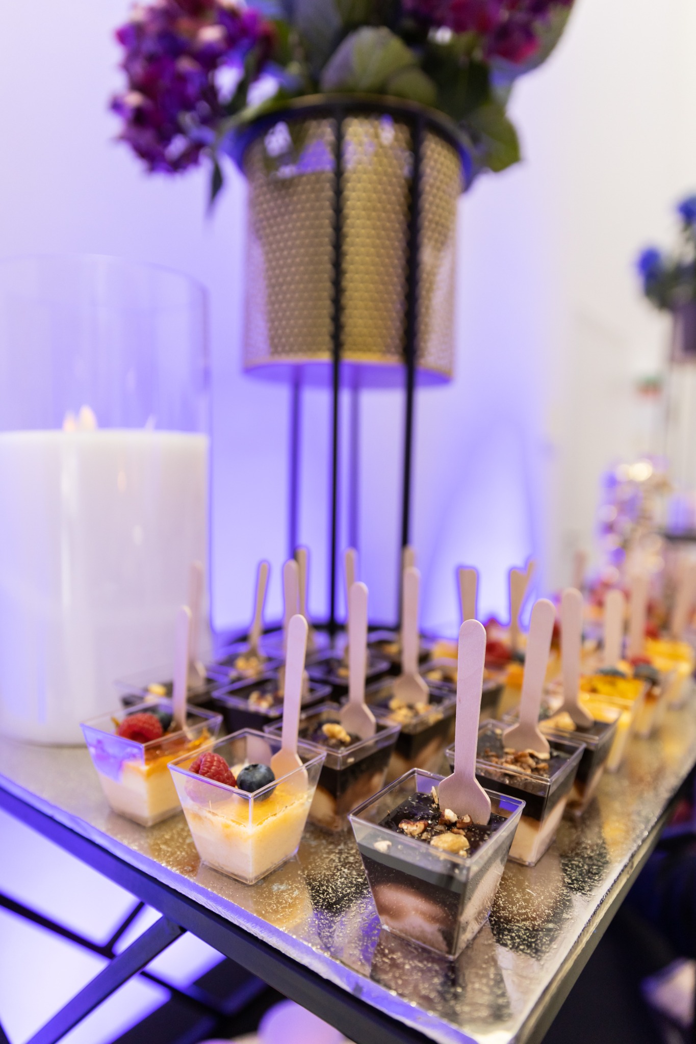 Deserki w małych plastikowych pojemnikach z drewnianymi łyżeczkami ustawione na srebrnym stole, w tle kwiaty w złotym wazonie i świeca, oświetlenie w odcieniach fioletu.
