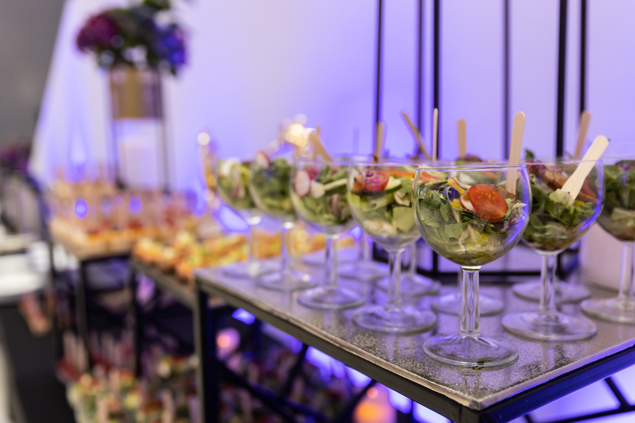 Szklanki z sałatkami na stole cateringowym, oświetlone fioletowym światłem, widoczne radyjki i pomidory koktajlowe.