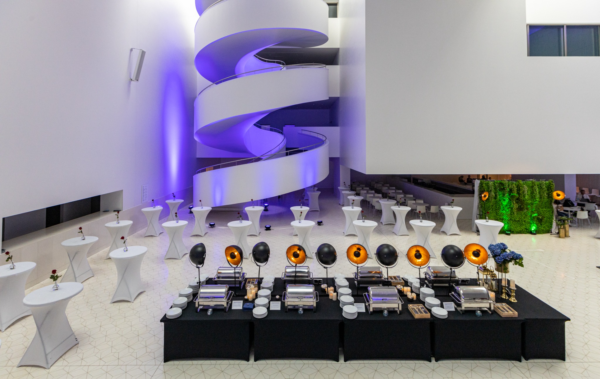 Elegancko przygotowana sala z bufetem, stolikami koktajlowymi z różami i spiralnymi schodami w tle, oświetlona fioletowym światłem.