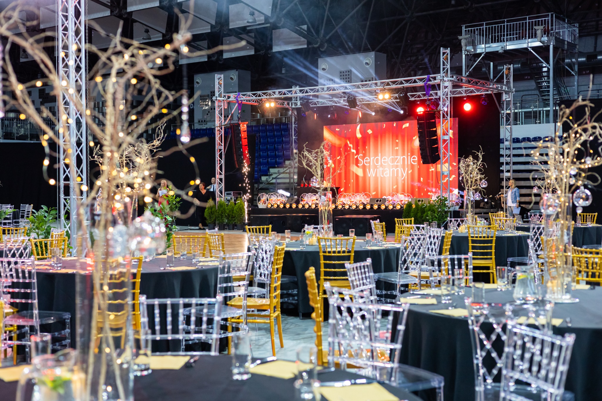Uroczysta sala bankietowa z okrągłymi stołami nakrytymi czarnymi obrusami, przezroczystymi i złotymi krzesłami, dekoracjami ze srebrnych gałązek z lampkami, sceną w tle z napisem 'Serdecznie...