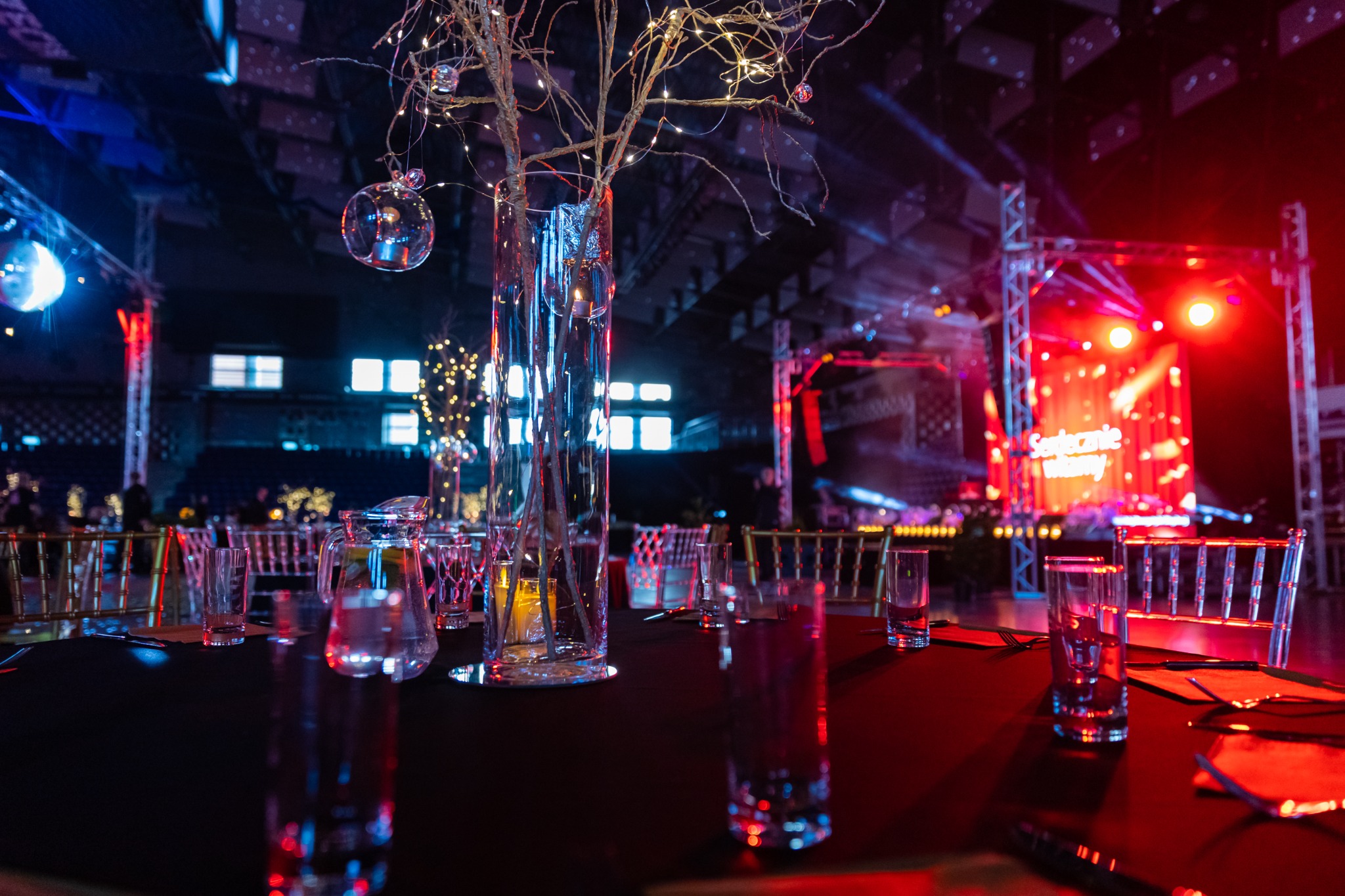 Uroczysta dekoracja stołu z wysokim wazonem z gałązkami i światełkami, szklanki i karafki, w tle scena z oświetleniem w kolorach czerwieni i błękitu, przygotowana na imprezę.