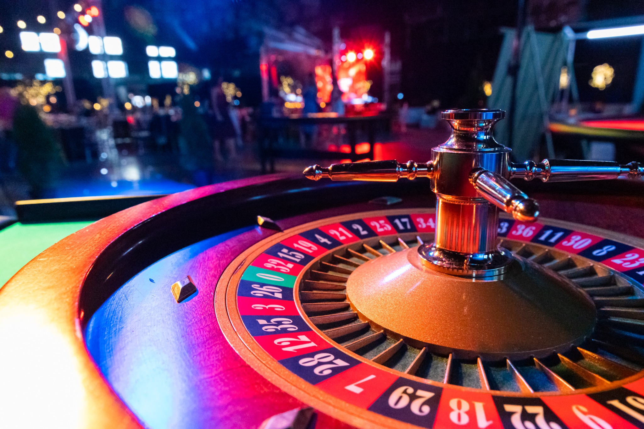 Roulette wheel with numbers and golden accents, illuminated by colorful stage lights in a casino setting, suggesting a high-stakes event.