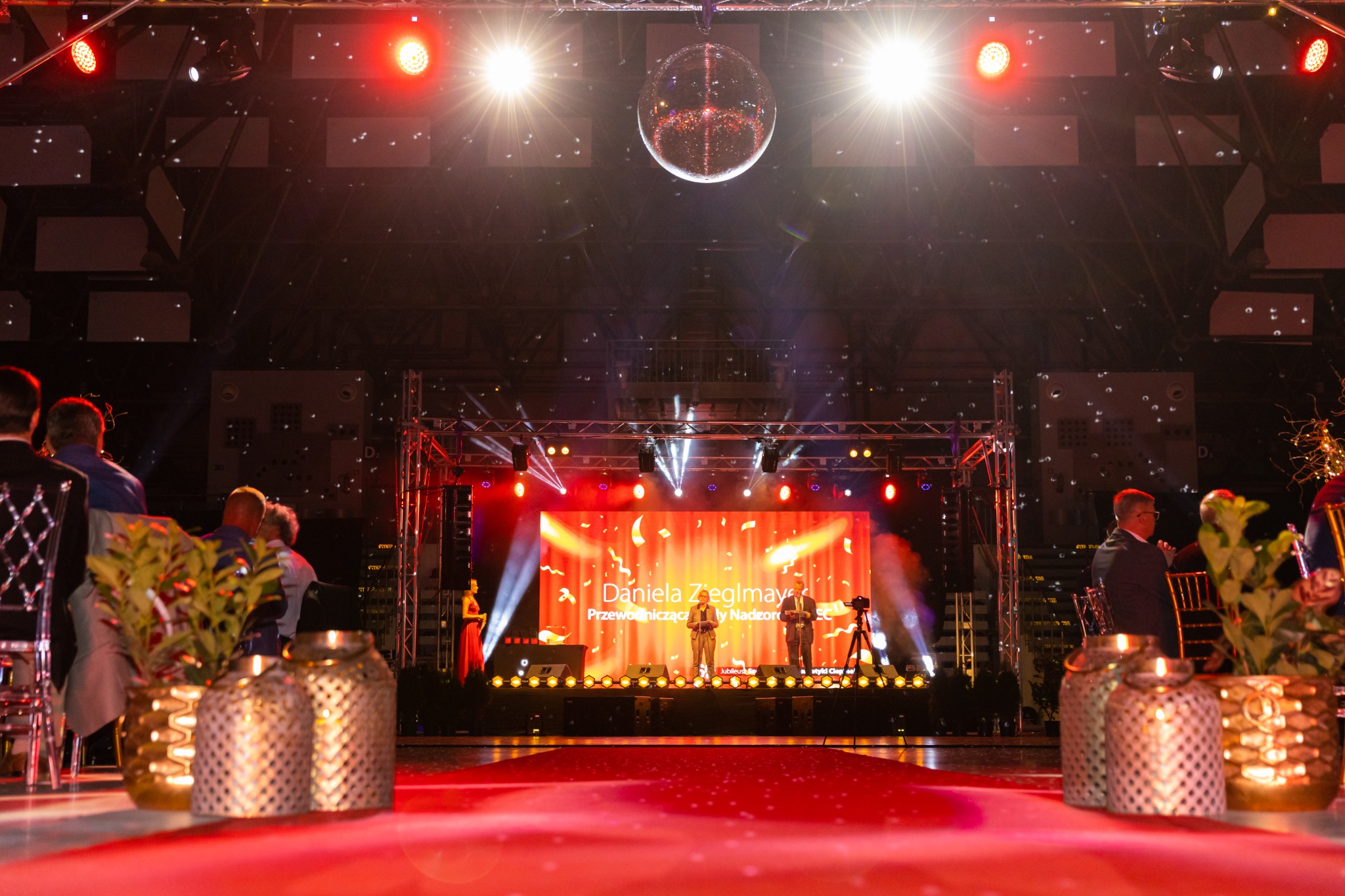 Scena podczas uroczystości z czerwonym dywanem, oświetleniem scenicznym, dekoracjami i ekranem wyświetlającym nazwisko prelegenta.
