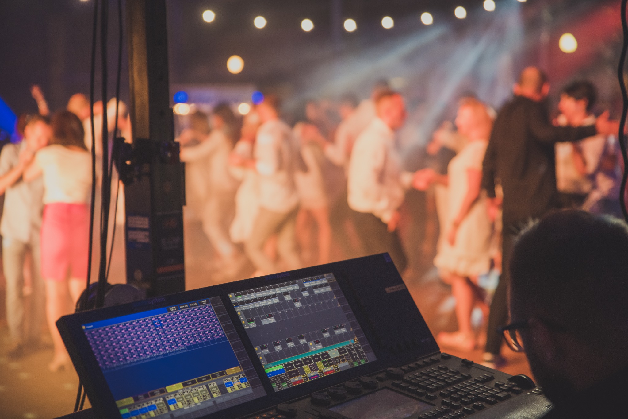Konsoleta oświetleniowa z widokiem na tańczących ludzi w tle, oświetlonych reflektorami podczas imprezy.
