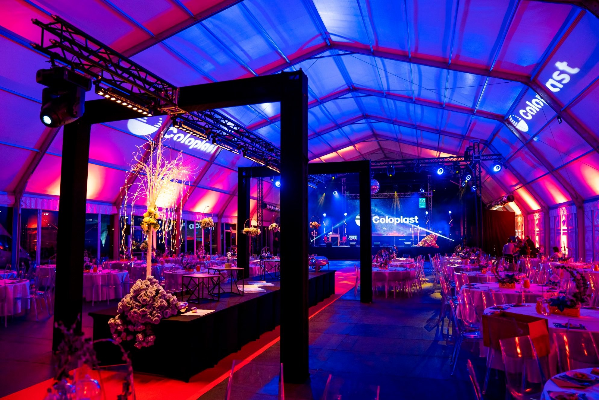 Uroczysta gala firmowa w namiocie z oświetleniem w odcieniach różu i błękitu, dekoracje stołów z kwiatami, scena z logo firmy Coloplast w tle.