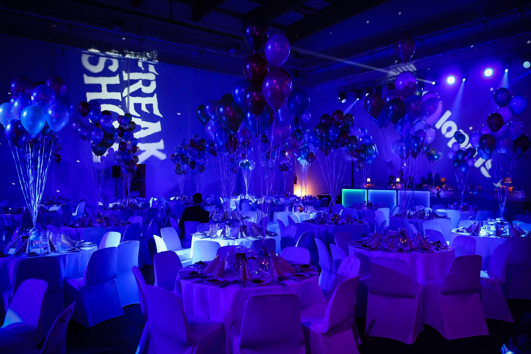 Uroczysta sala bankietowa oświetlona na niebiesko z okrągłymi stołami nakrytymi białymi obrusami i krzesłami w pokrowcach, udekorowana balonami, z projekcjami świetlnymi na ścianach.