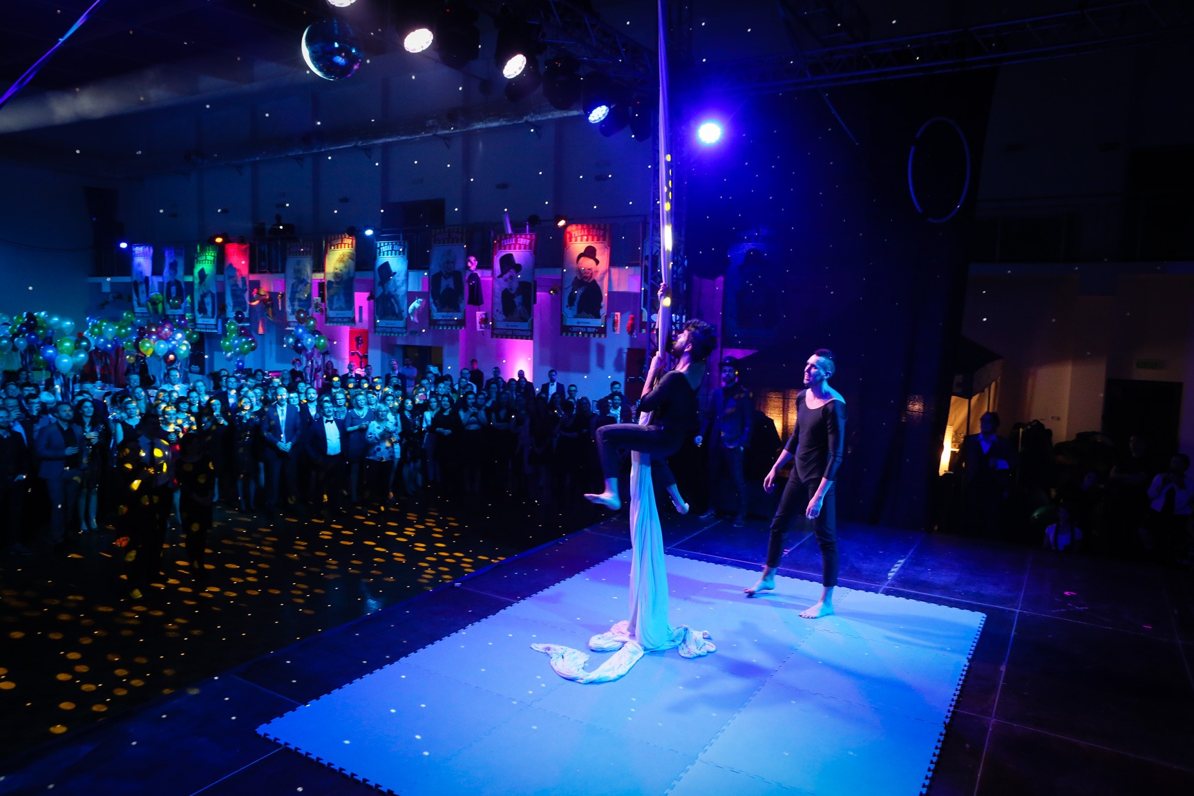 Dwóch artystów prezentuje akrobatyczny występ na szarfie podczas imprezy firmowej oświetlonej niebieskim światłem, z widoczną publicznością w tle i dekoracjami tematycznymi.