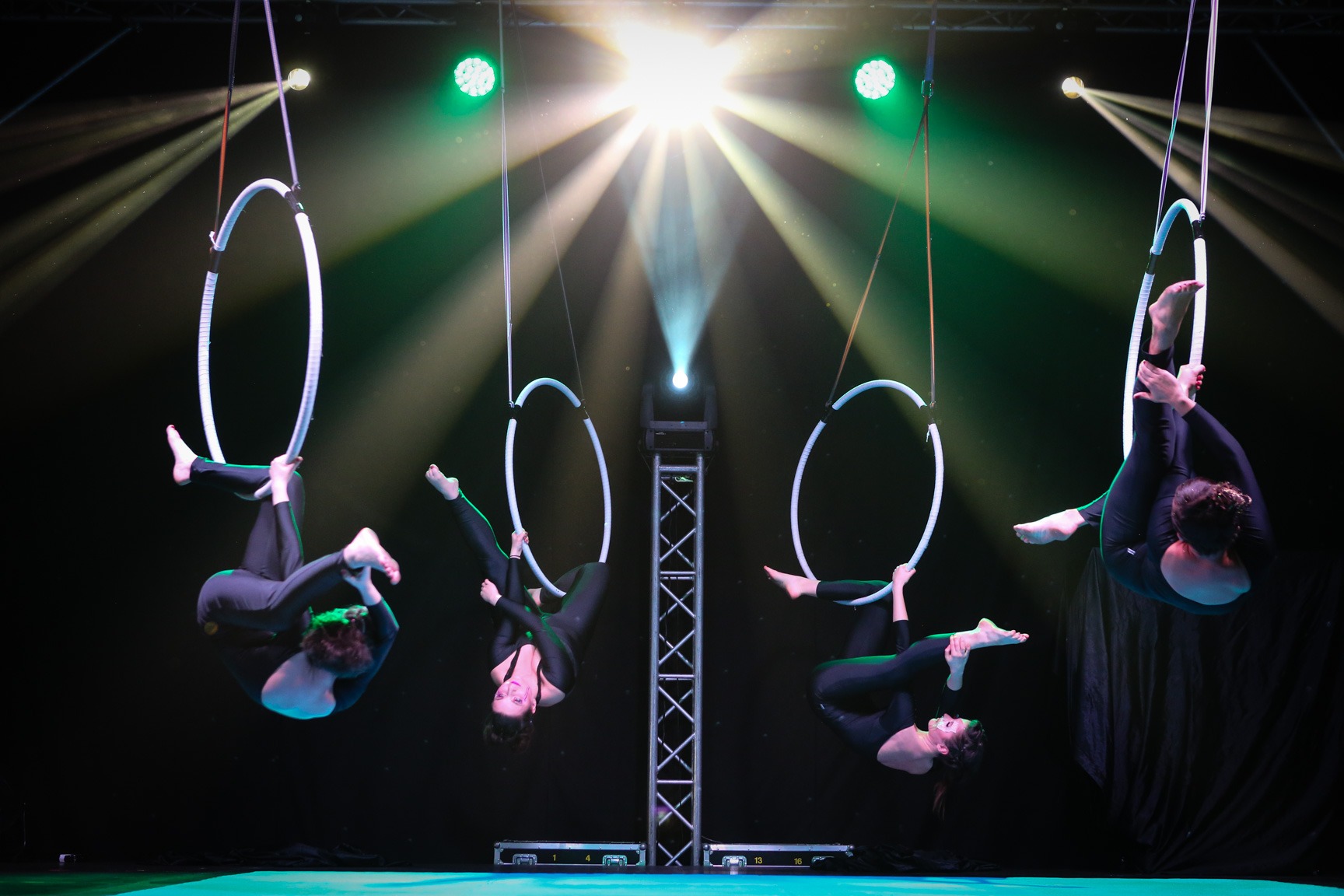 Cztery kobiety w czarnych kostiumach wykonują akrobacje powietrzne na metalowych obręczach zawieszonych pod sceną, oświetlone reflektorami w kolorach białym i zielonym.