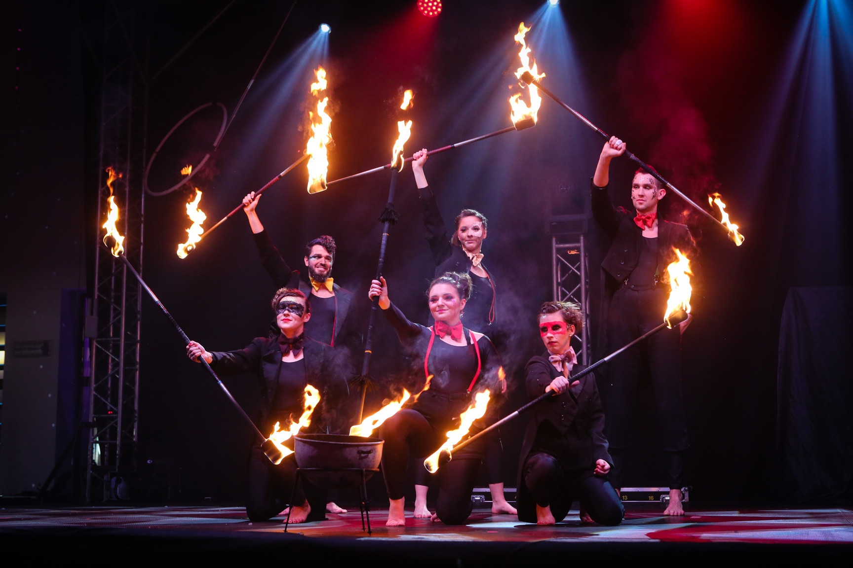 Grupa artystów w czarnych strojach z muszkami i maskach prezentuje pokaz z płonącymi pochodniami na scenie oświetlonej reflektorami.
