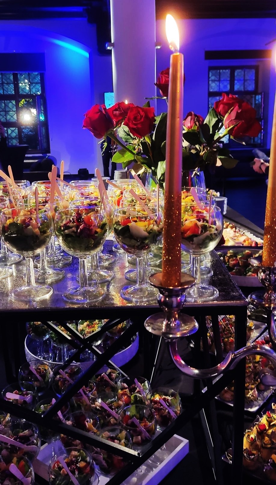 Elegancki bufet z sałatkami w kieliszkach, dekorowany świecami i czerwonymi różami, w tle niebieskie oświetlenie i okno.