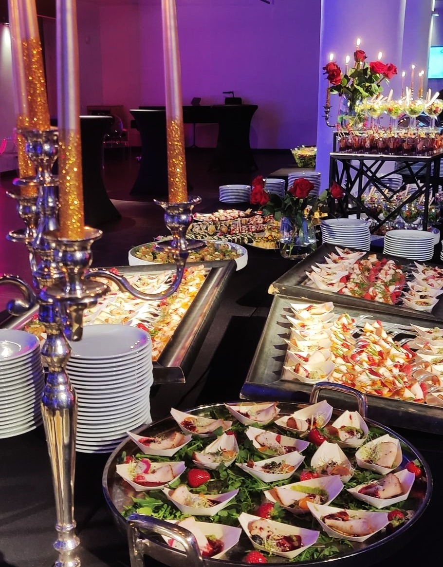 Bogato zastawiony stół z przekąskami na przyjęciu: srebrny świecznik z brokatowymi świecami, tace z różnorodnymi przystawkami, róże w wazonie i deserki w szklankach na metalowym stojaku.