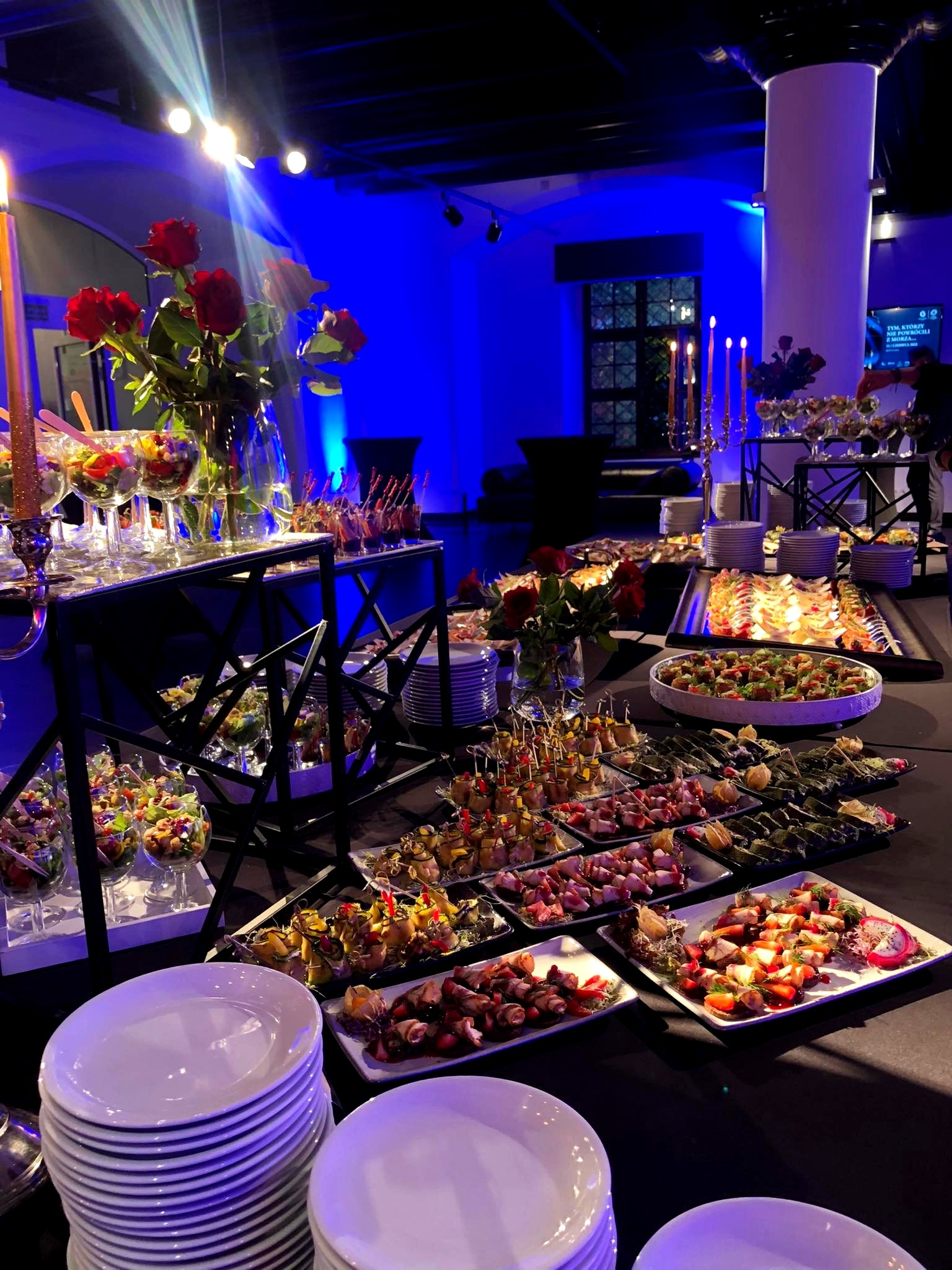 Bogato zastawiony stół z przekąskami i dekoracjami kwiatowymi w sali z niebieskim oświetleniem, widoczne szklanki z sałatkami, koreczki i talerze z roladkami.