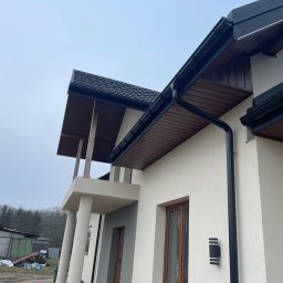 Fred Construction - Elewacja domu jednorodzinnego z widocznymi elementami wykończeniowymi: rynny, okna w drewnianej ramie, kolumny podtrzymujące zadaszenie balkonu, zewnętrzna lampa ścienna.