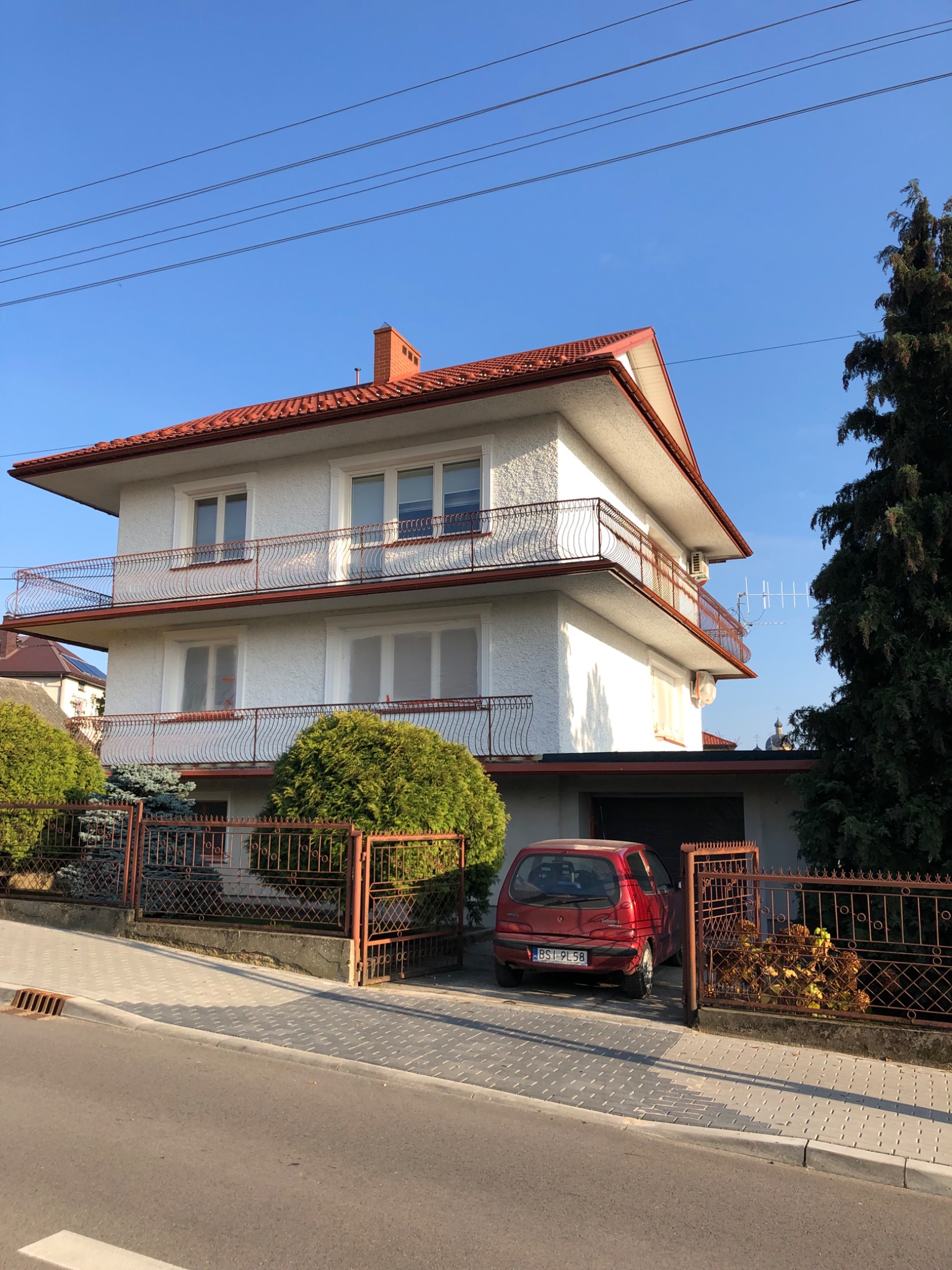Dwupiętrowy dom z białym tynkiem i czerwonym dachem, widoczne balkony z metalowymi balustradami, okna częściowo zakryte folią malarską, zaparkowany czerwony samochód przy bramie.