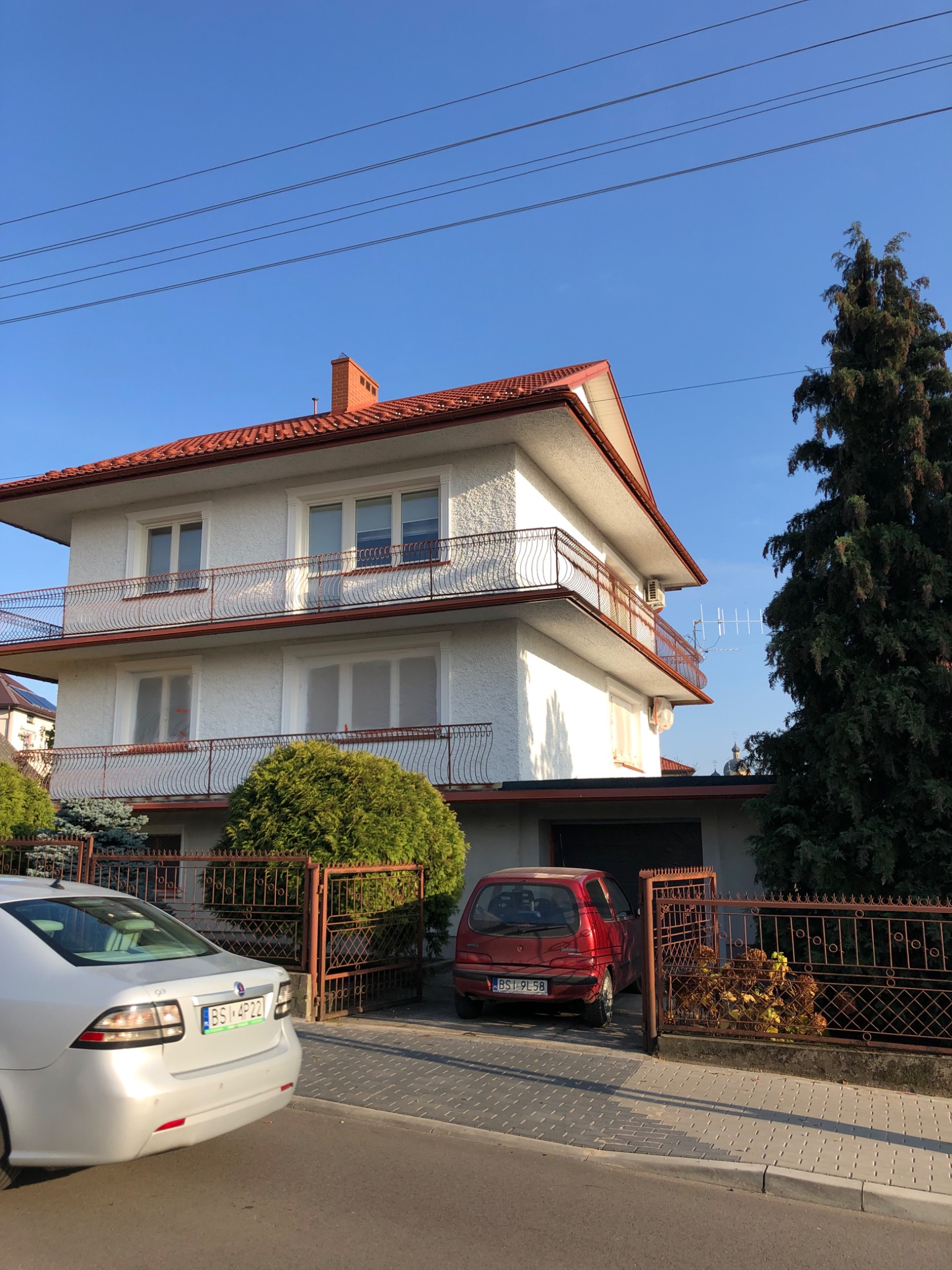 Dwupiętrowy dom z białymi ścianami i czerwoną dachówką, widoczne balkony z metalowymi balustradami, samochody na podjeździe, brama z metaloplastyki.