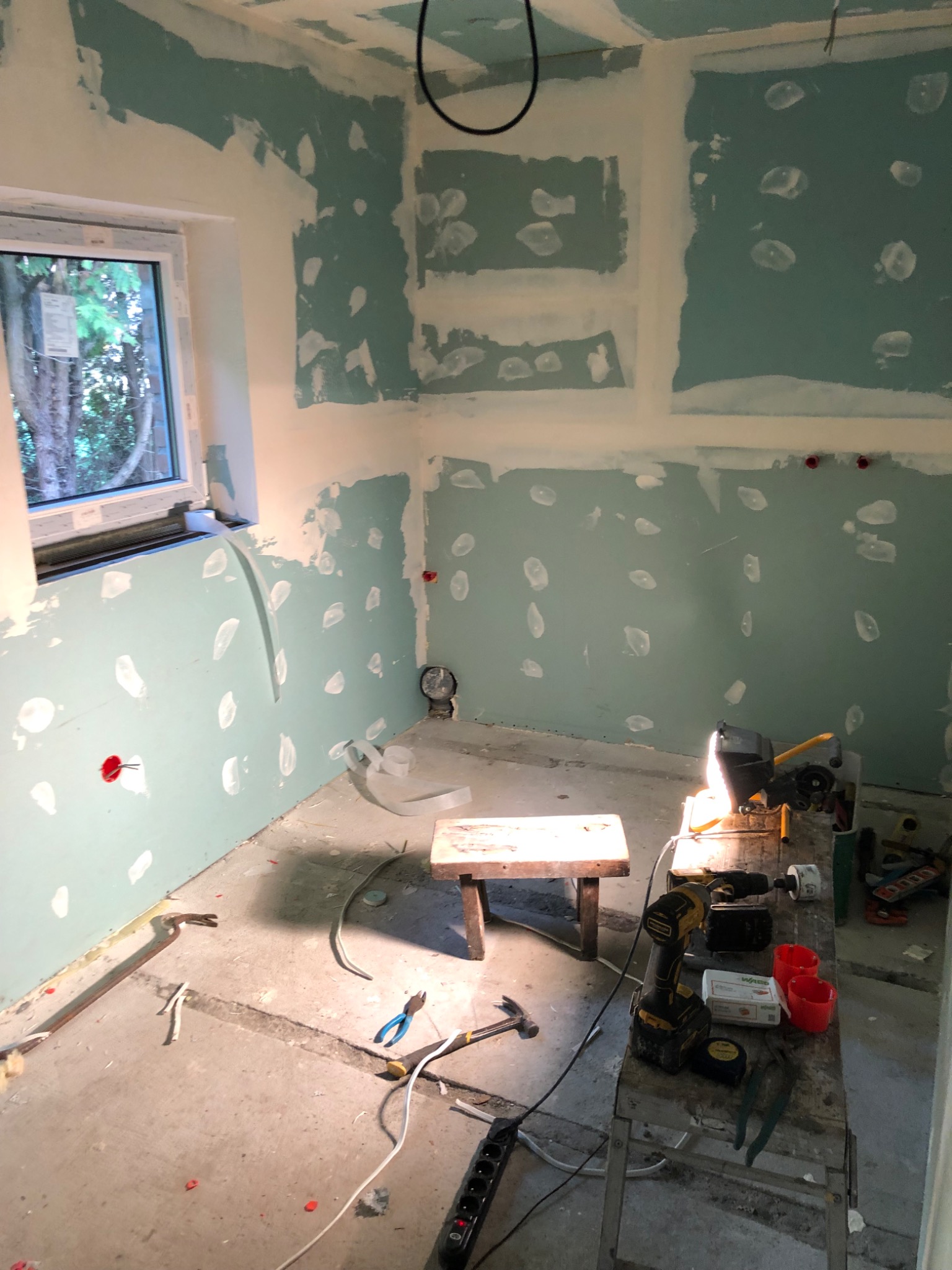 Pokój w trakcie remontu: ściany z płyt kartonowo-gipsowych z widocznymi punktami szpachlowania, narzędzia i materiały na metalowym stole warsztatowym, mały drewniany stołek pośrodku, okno...