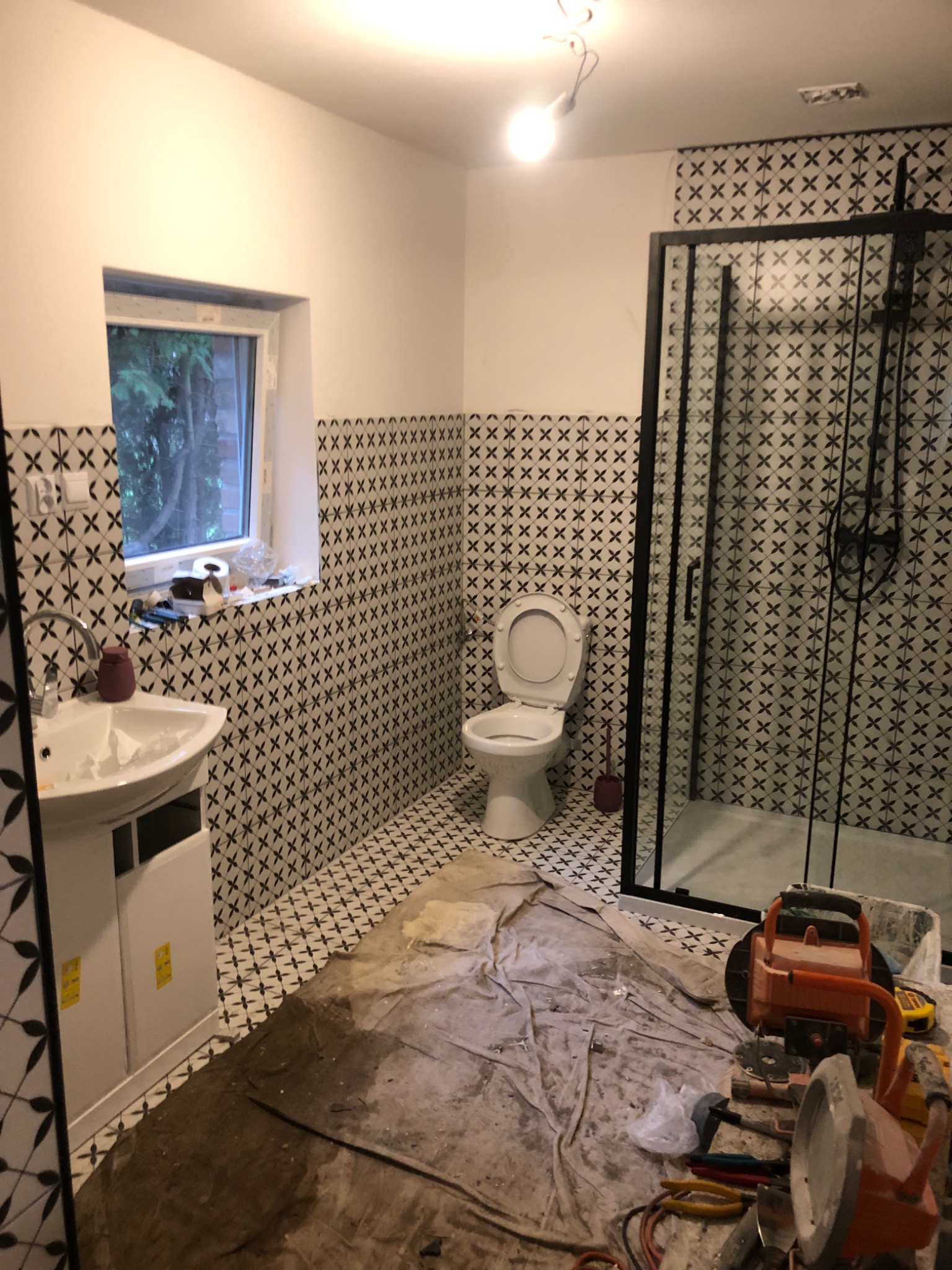 Łazienka w trakcie remontu: białe ściany z czarnym wzorem, kabina prysznicowa z czarną ramą, toaleta, umywalka, podłoga zabezpieczona folią, widoczne narzędzia budowlane.