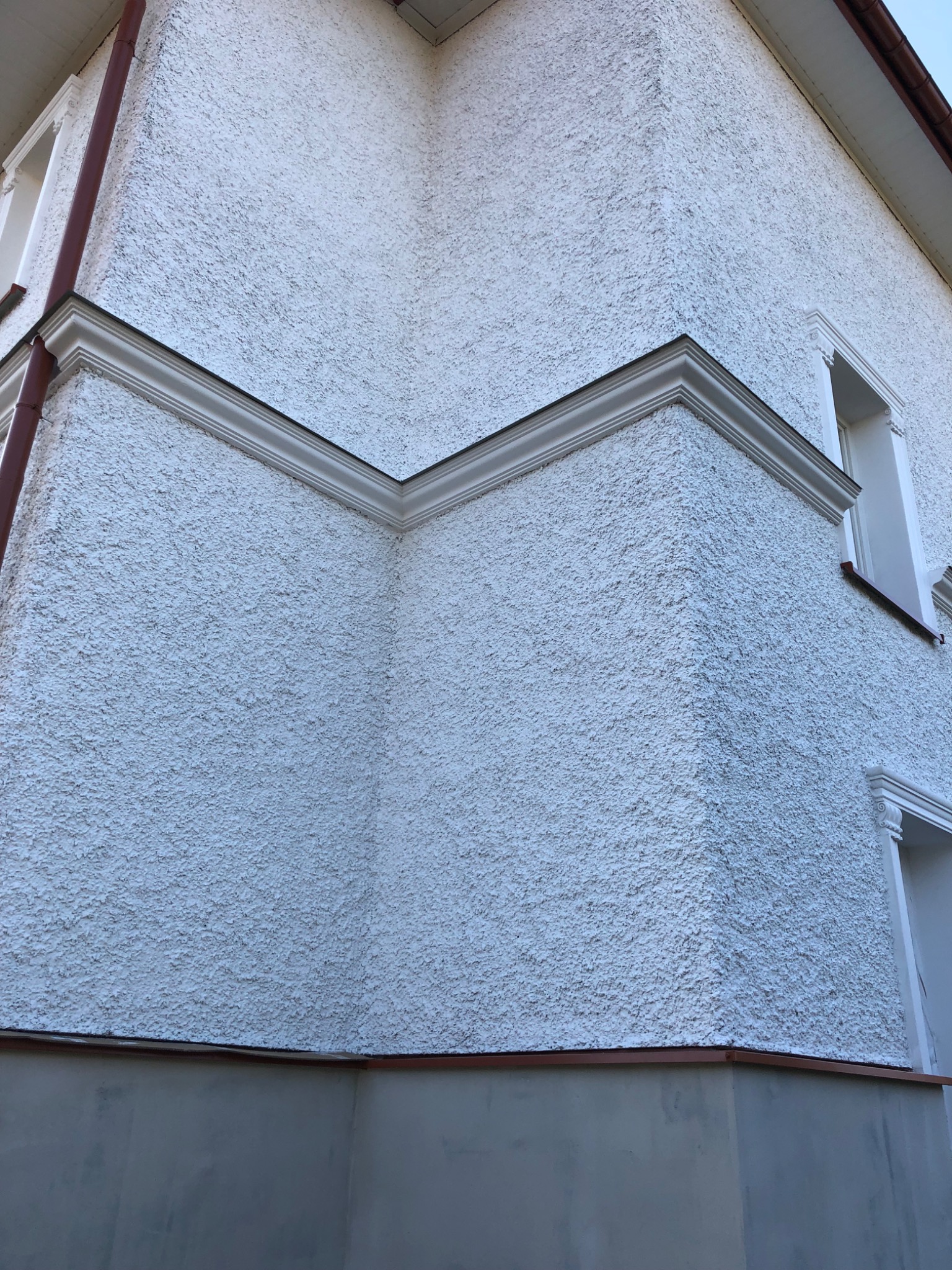 Narożnik budynku z białą, chropowatą elewacją, ozdobiony białym gzymsem i detalami okiennymi, uchwycony z perspektywy żabiej z lekkim pochyleniem.