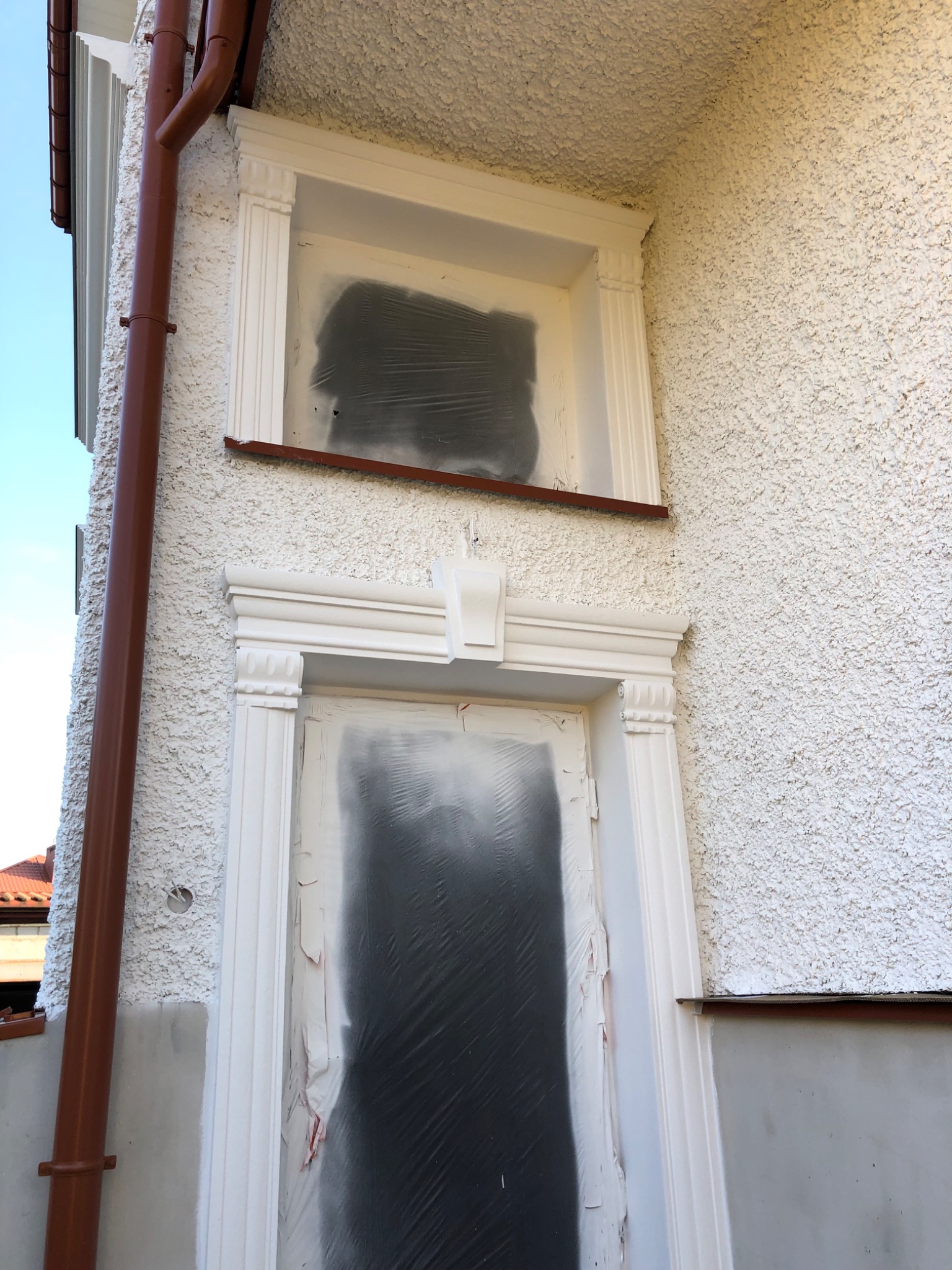 Elewacja budynku z oknem i drzwiami zabezpieczonymi folią malarską przed świeżo nałożonym tynkiem strukturalnym, widoczna rynna.