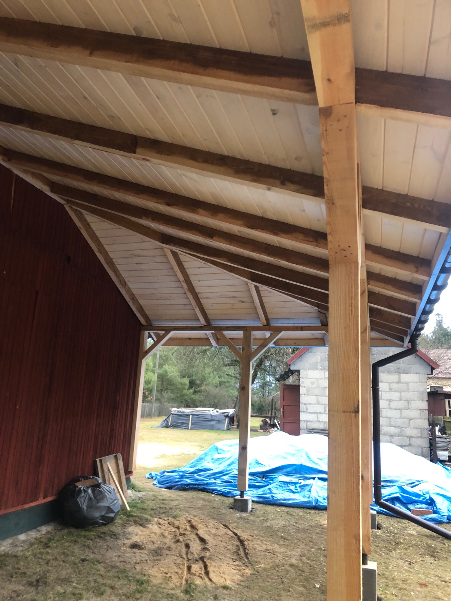 Nowa drewniana konstrukcja z białym podbitką dachową, widoczne krokwie i słupy nośne, w tle budynki i teren budowy zabezpieczony niebieską folią.