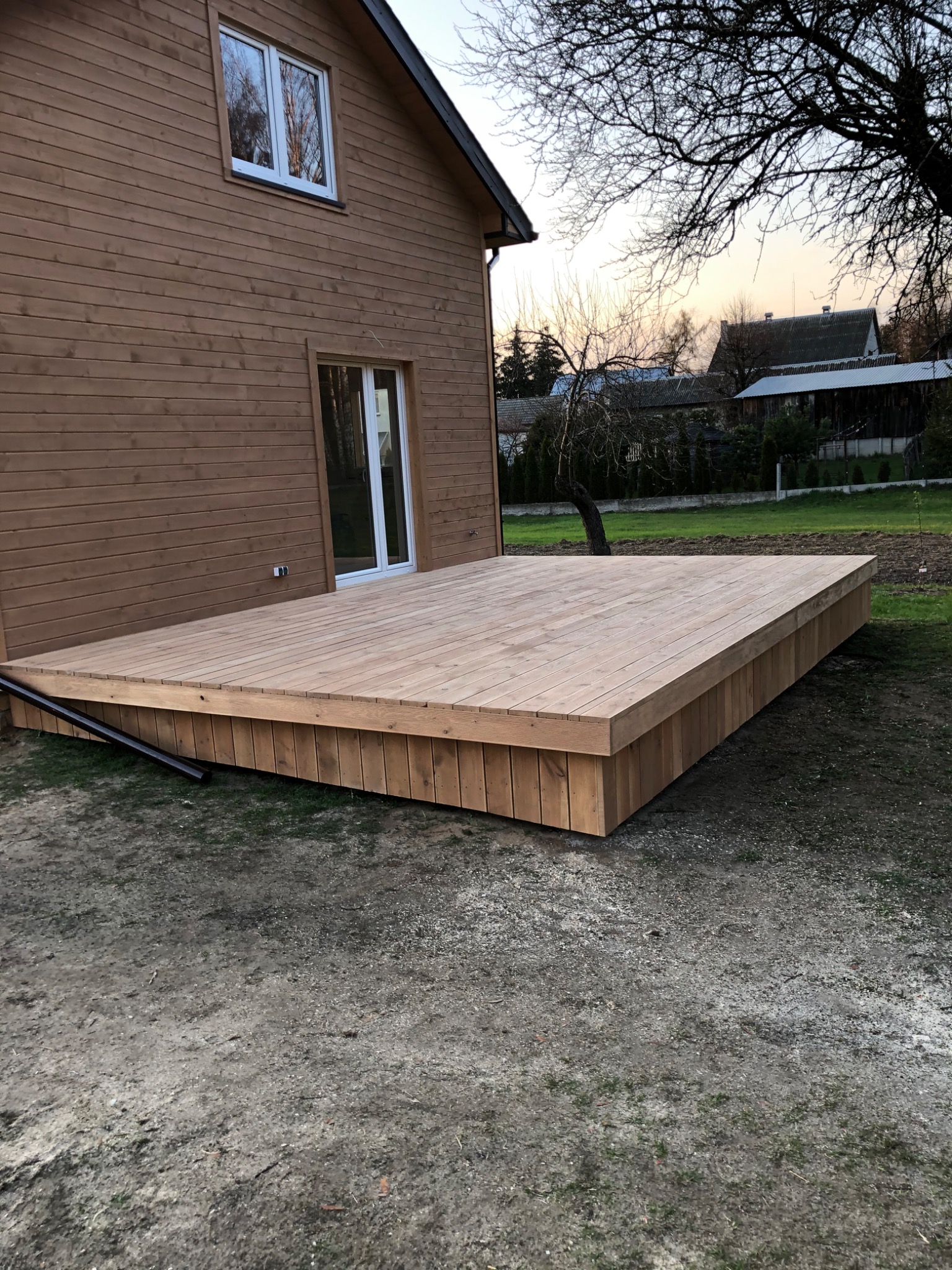 Drewniany taras przylegający do domu z drewnianą elewacją, widok z poziomu gruntu, rynna odprowadzająca wodę z dachu widoczna przy tarasie.