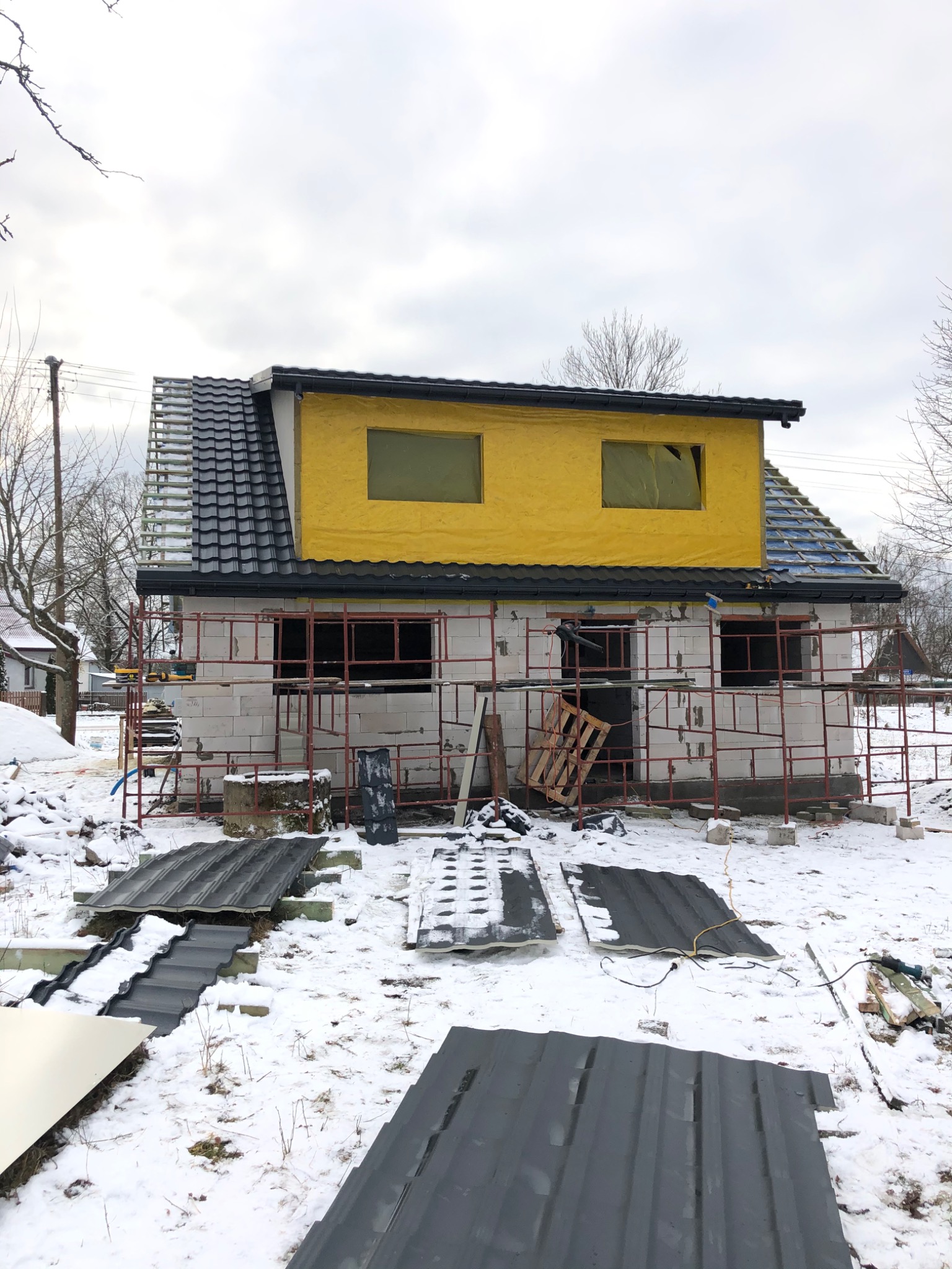 Dom w trakcie budowy zimą, z widocznym rusztowaniem, częściowo położonym dachem z czarnej blachy, oknami zabezpieczonymi żółtą folią i stertą arkuszy blachy na śniegu.